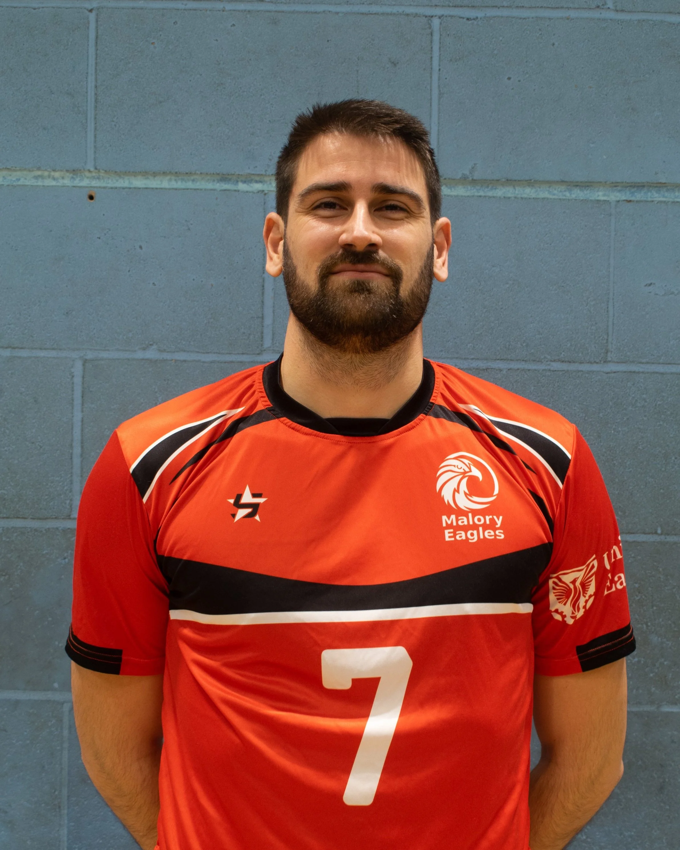 A man with dark hair and a beard in a red volleyball jersey with the number 7, standing against a blue brick wall.