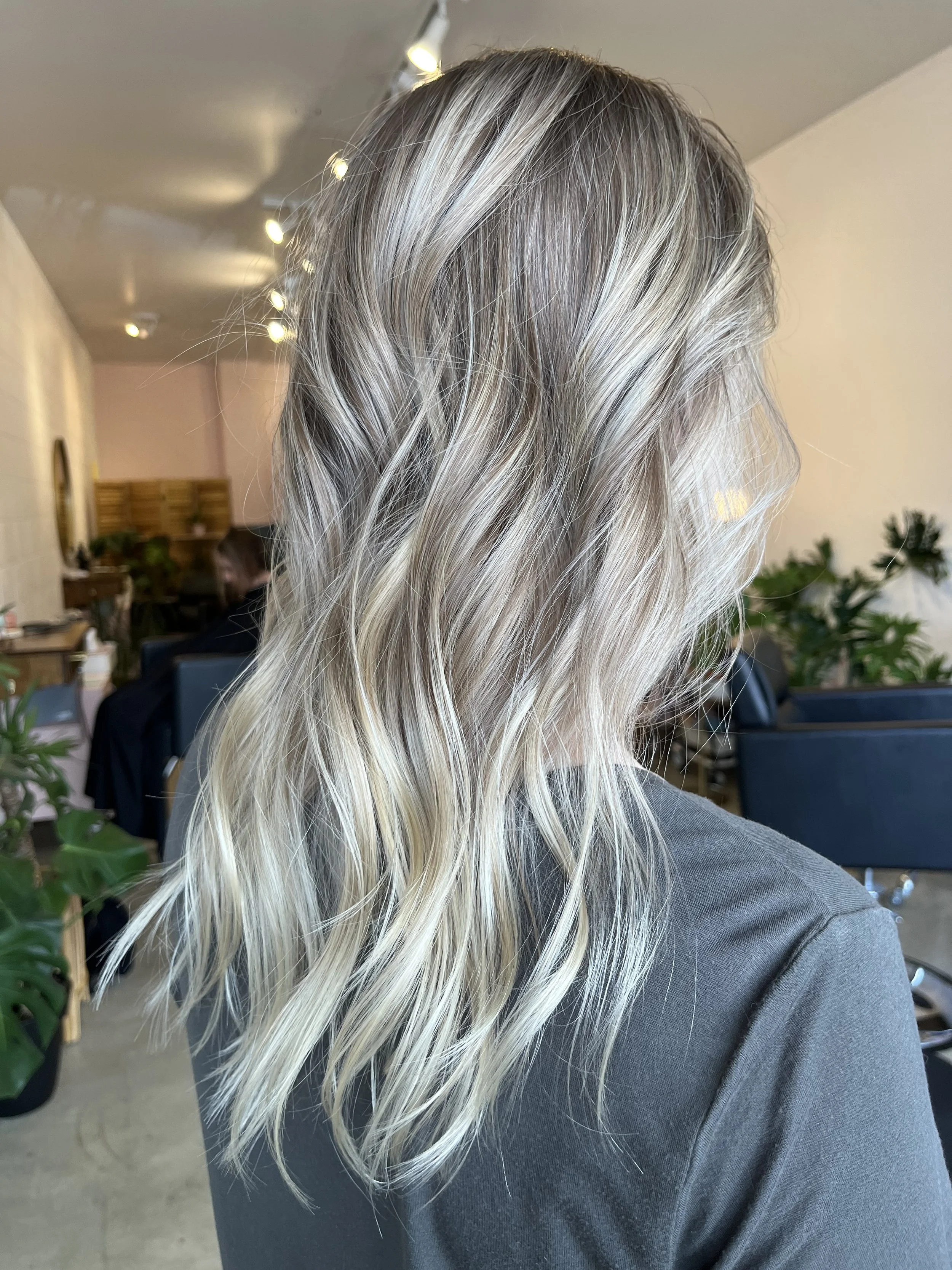 Side view of a woman with long, wavy blonde hair sitting indoors with plants and furniture visible in the background.