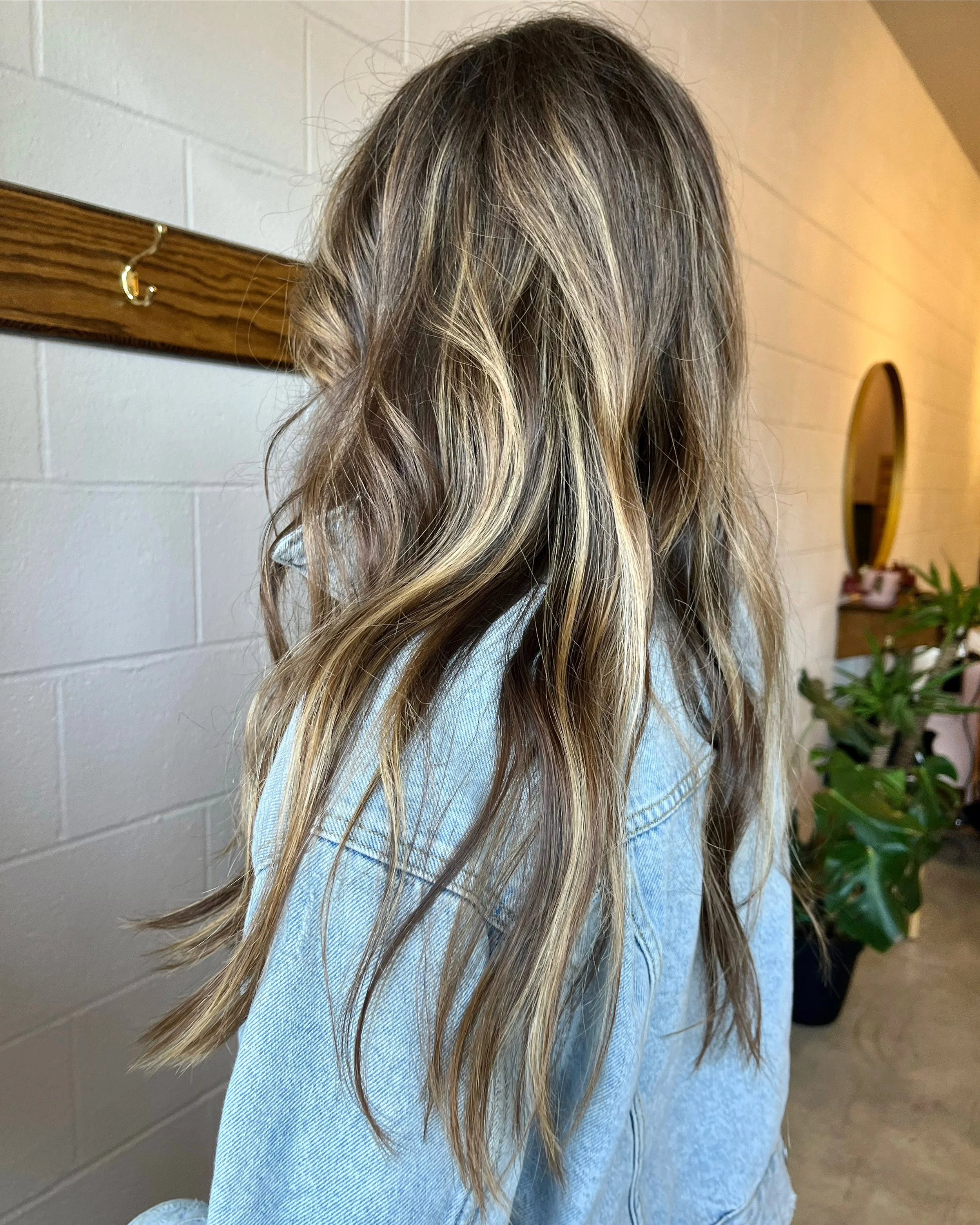 Back view of a person with long, wavy, blonde hair wearing a light blue denim jacket in a room with a white wall, wooden décor, a mirror, and potted plants.