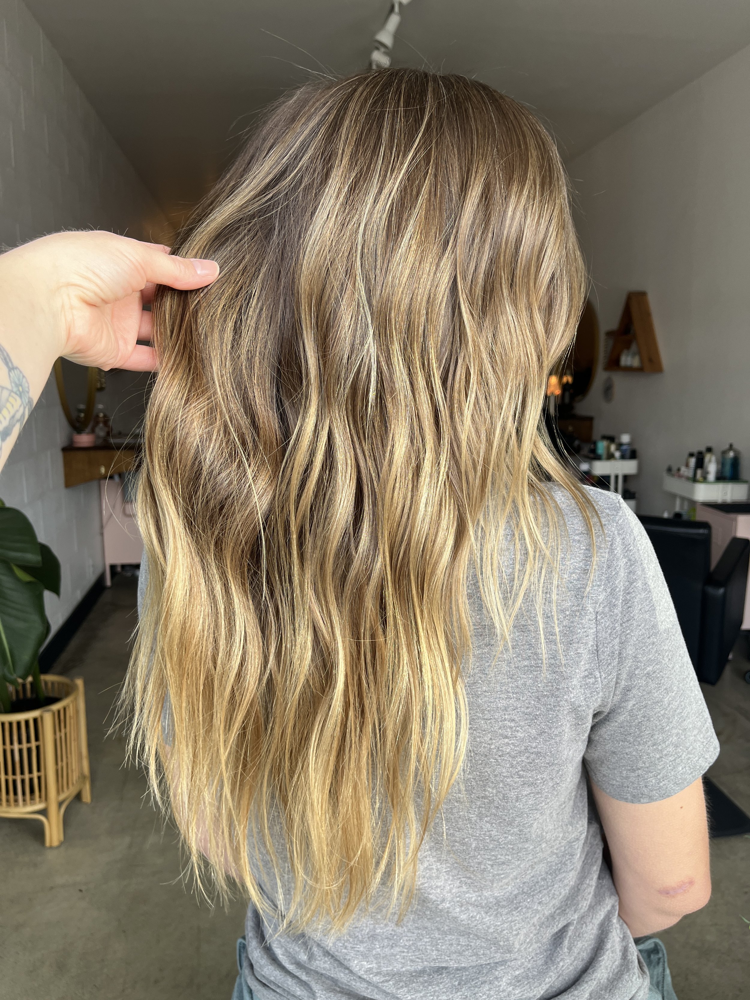 Back of a woman with wavy blonde hair wearing a gray shirt inside a hair salon.