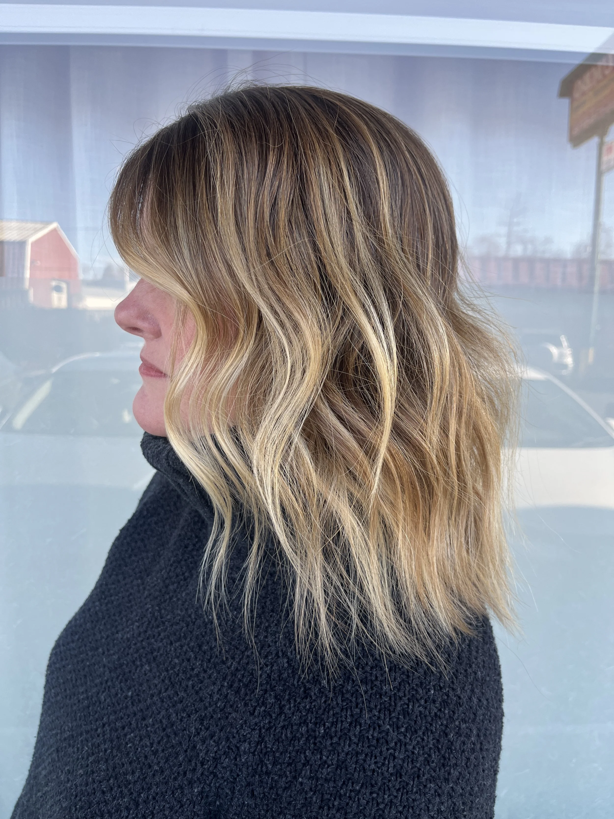 Woman with wavy, shoulder-length blonde hair and darker roots, wearing a black sweater, standing in front of a window with a blurred outdoor scene.