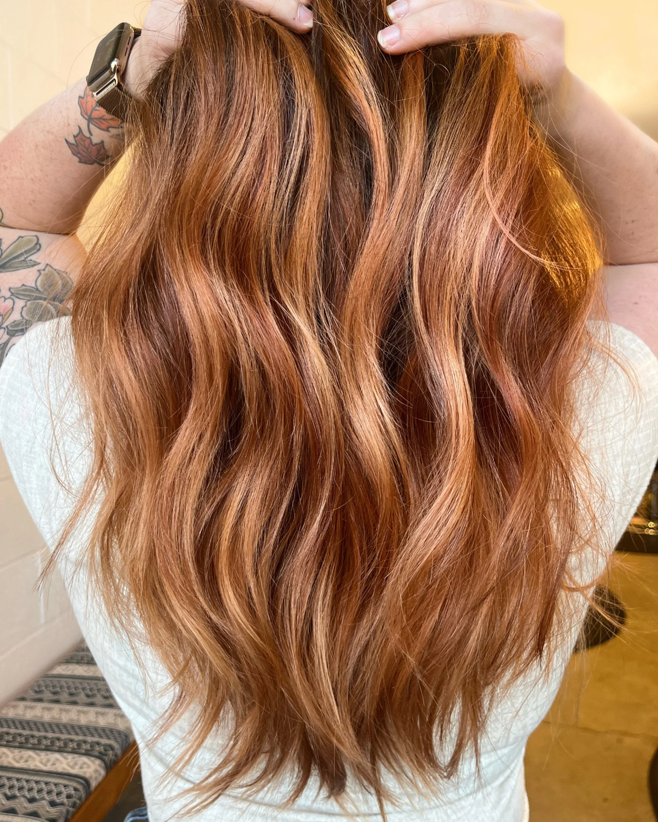 Close-up of a person with long, wavy, reddish-brown hair and tattoos on their arms, holding their hair up with both hands.