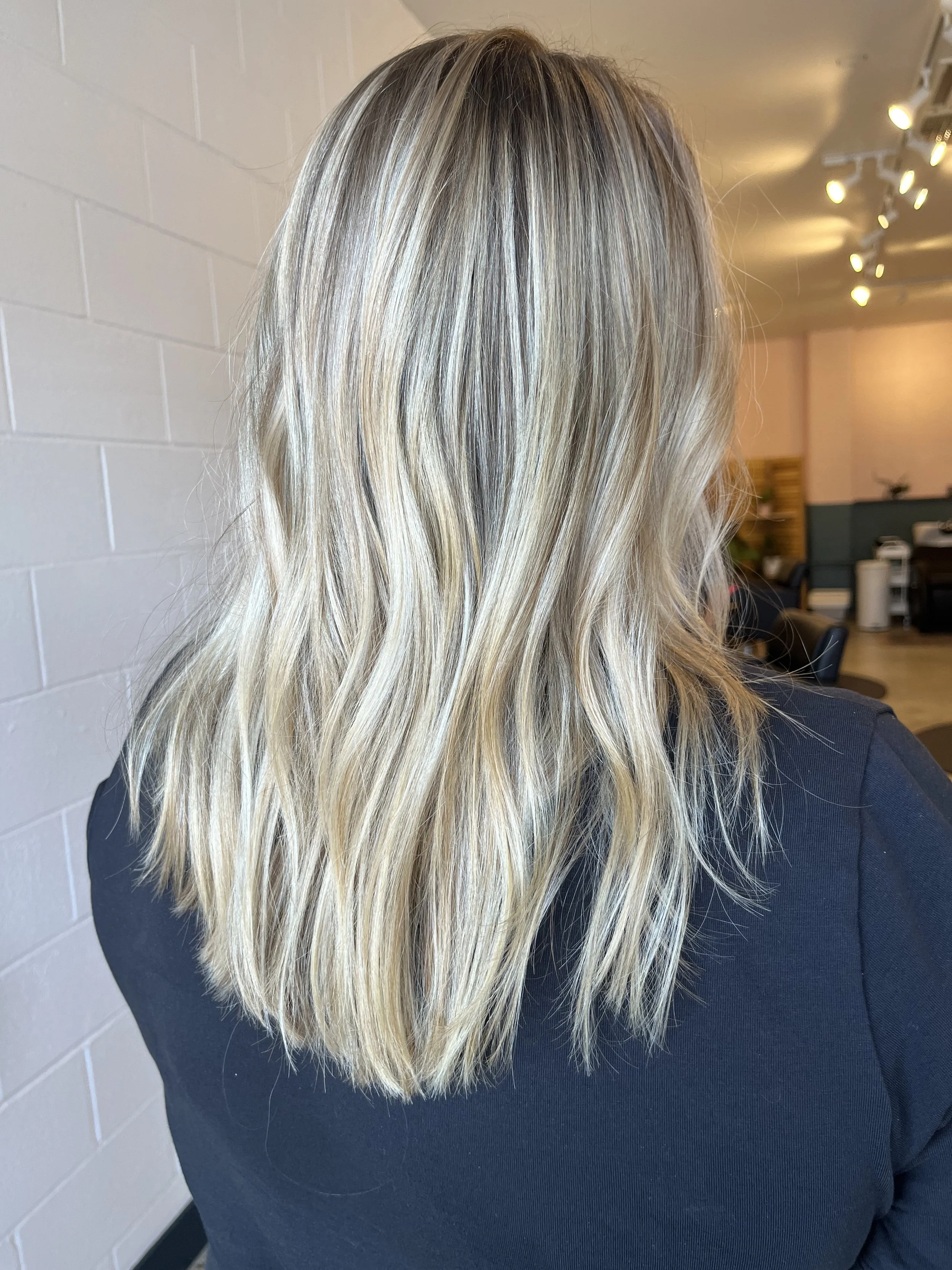 Back view of a woman with long, wavy blonde hair, wearing a black top, indoors with a white brick wall and lighting fixture in the background.