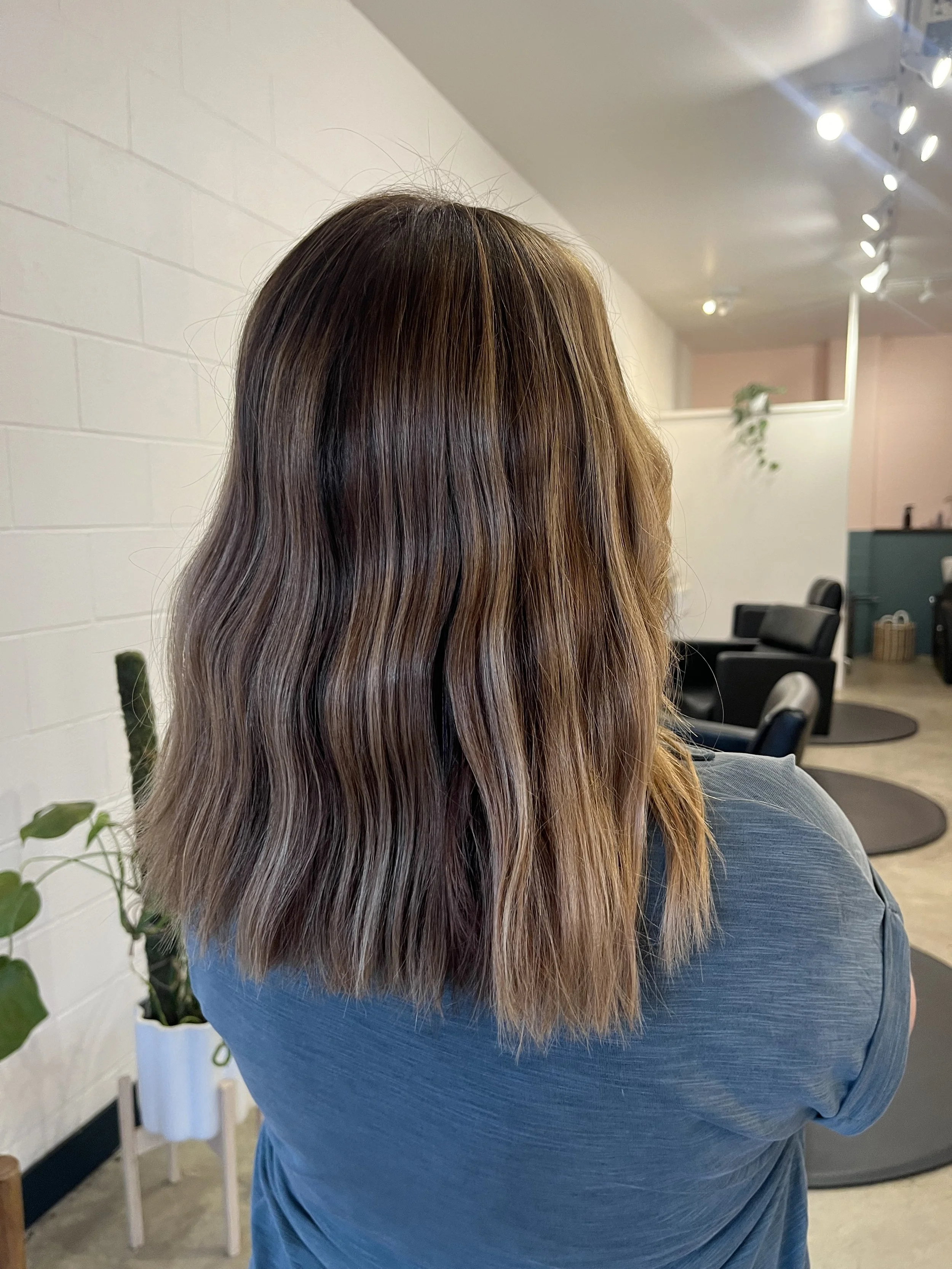 Back of a woman with shoulder-length wavy brown hair with blonde highlights, standing in a modern salon with a white brick wall and black chairs in the background.
