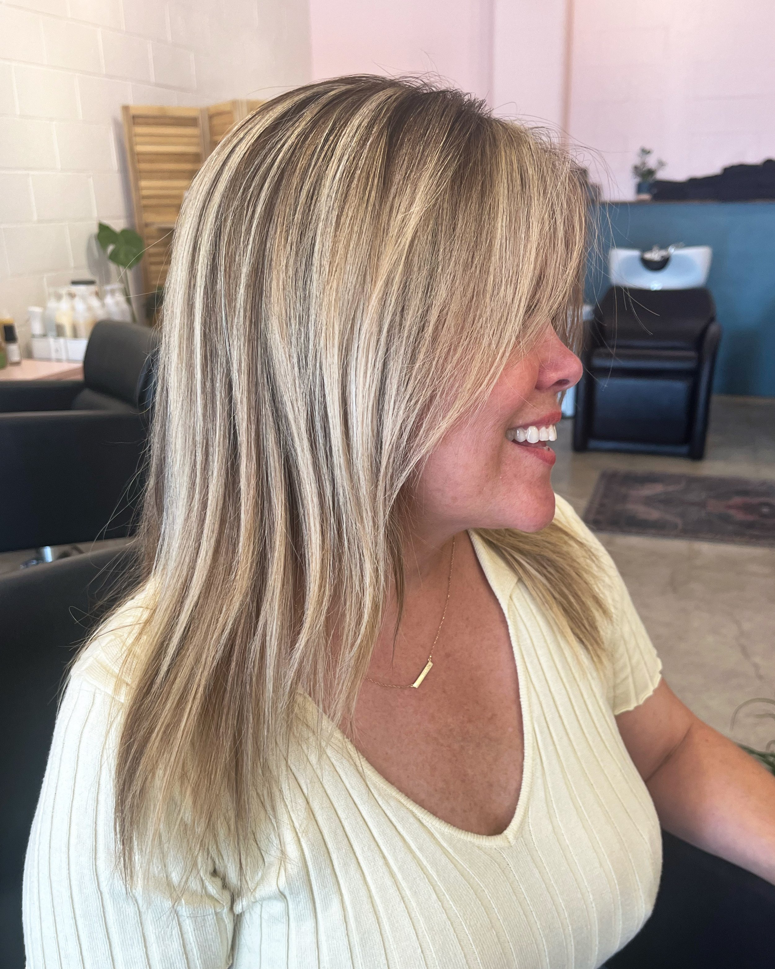 A woman with blonde hair sitting in a salon chair, smiling, wearing a cream-colored top and a gold necklace, with salon equipment and decor in the background.