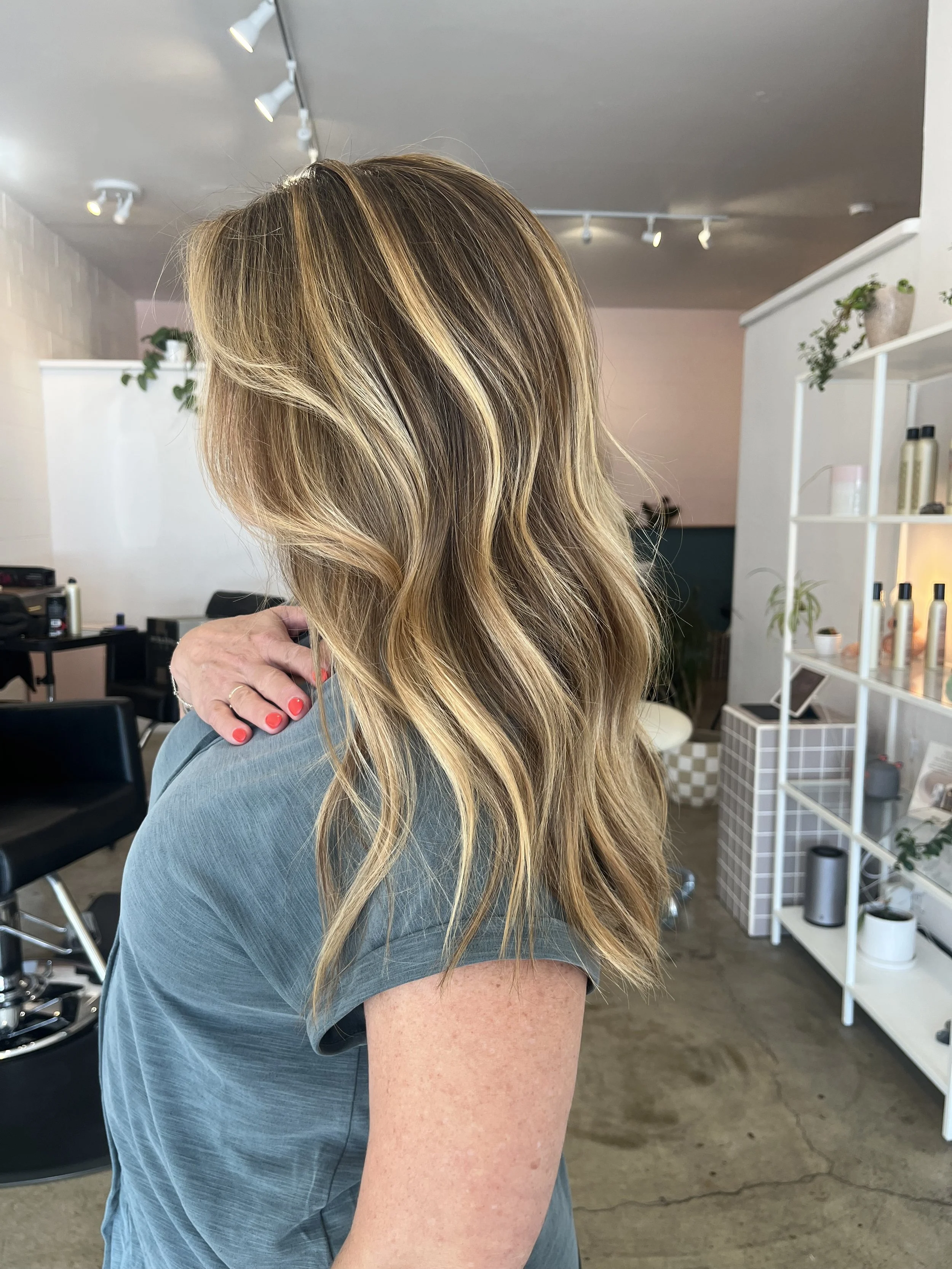 Side view of a woman with long, wavy blonde hair, wearing a blue-gray shirt, inside a modern salon with plants and shelves in the background.