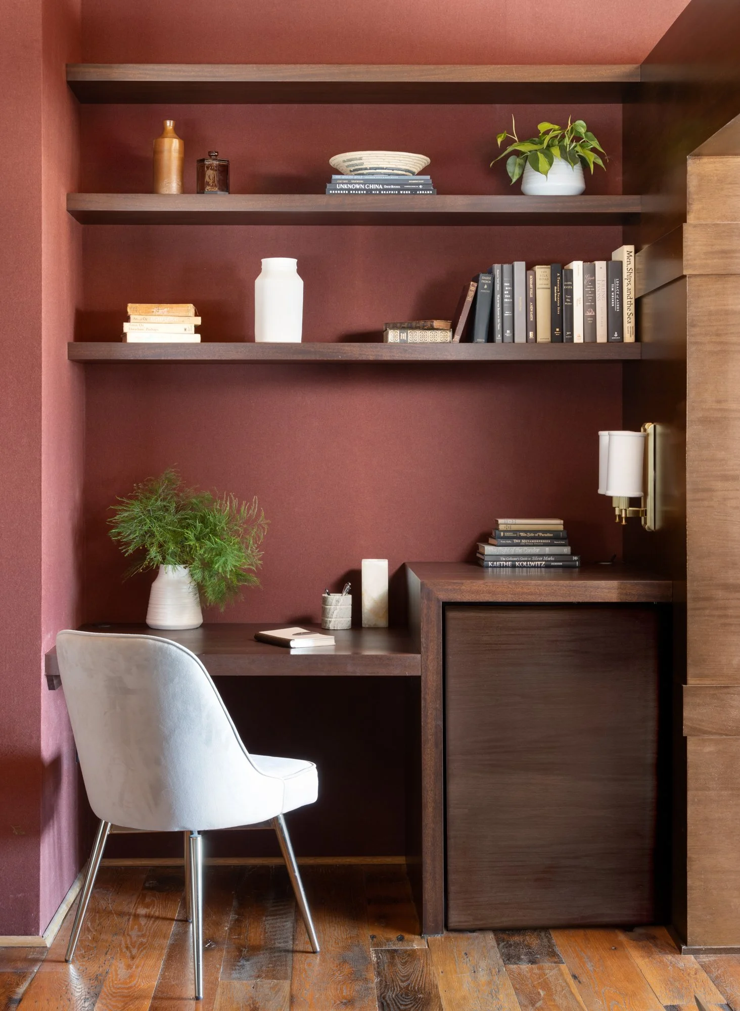 A cozy home office corner with a white chair, wooden desk, and burgundy wall with open shelves holding books, vases, and decorative items.