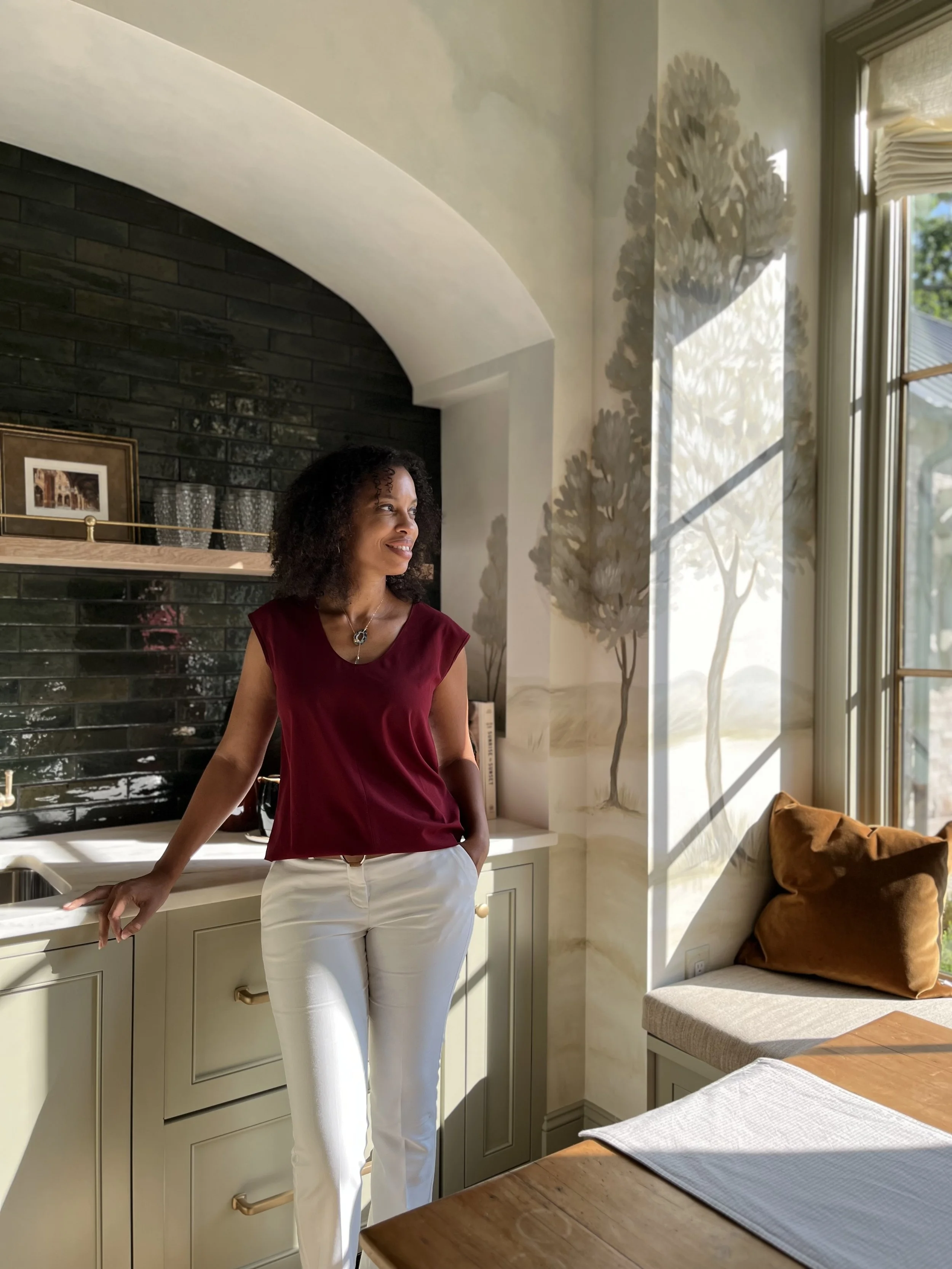 Laura Hodges in a burgundy top and white pants standing in a bright kitchen near a sink with sunlight streaming in and casting shadows on the wall with painted trees.