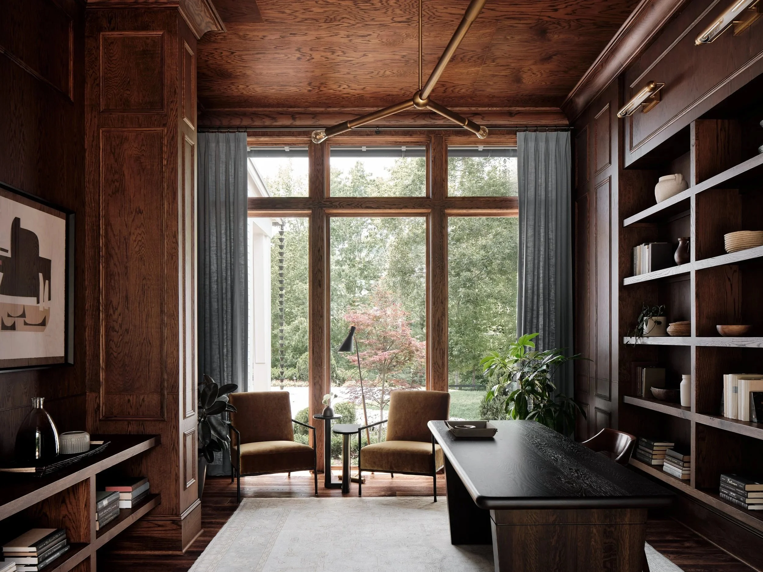 A cozy home office with dark wooden walls, a large window overlooking greenery, a black desk, two mustard chairs, bookshelf, plants, and a bookshelf with decorative items.