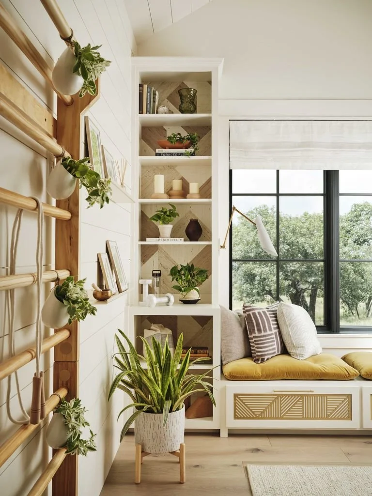 A cozy living room corner with a window seat cushion, built-in bookshelf with decorative items, potted plants, and a window showing lush greenery outside.