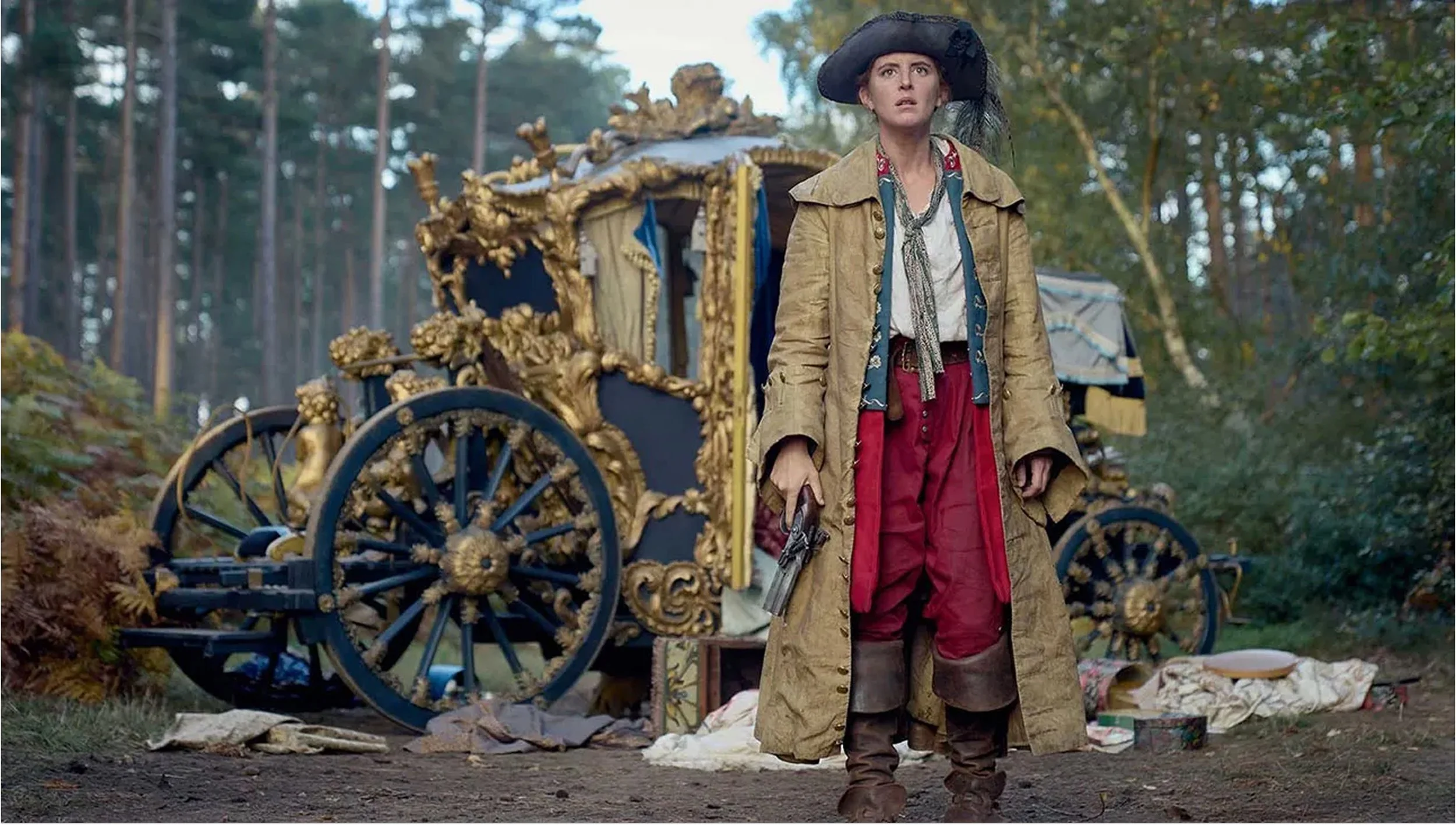 A woman dressed in 18th-century clothing, wearing a large black hat, holding a pistol, standing in front of an ornate, gold-trimmed carriage in a wooded area.