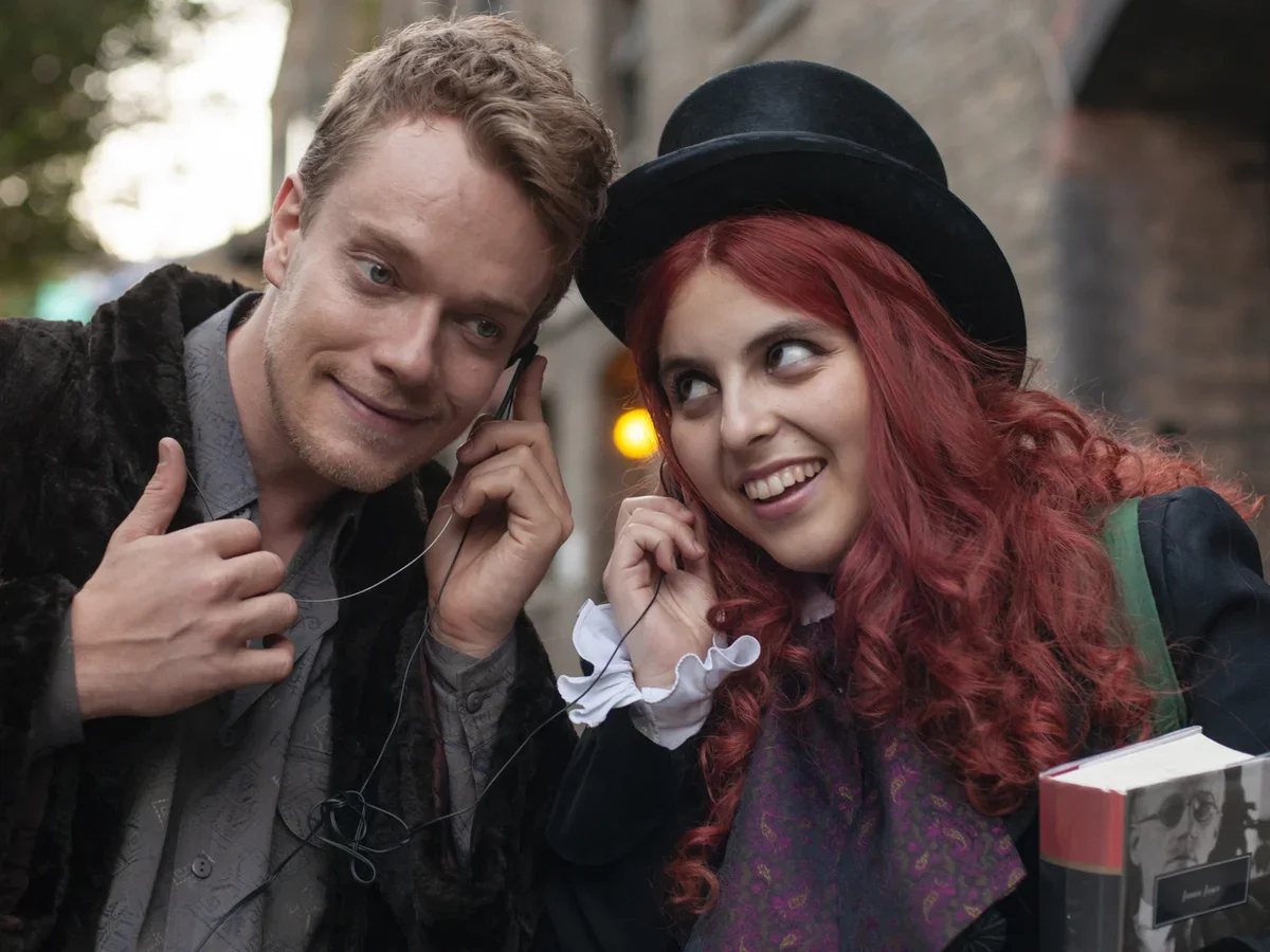 A young man and woman standing outdoors, both wearing headphones. The man is talking on a cellphone and giving a thumbs-up, while the woman, with red curly hair and a black top hat, is smiling and holding a book.