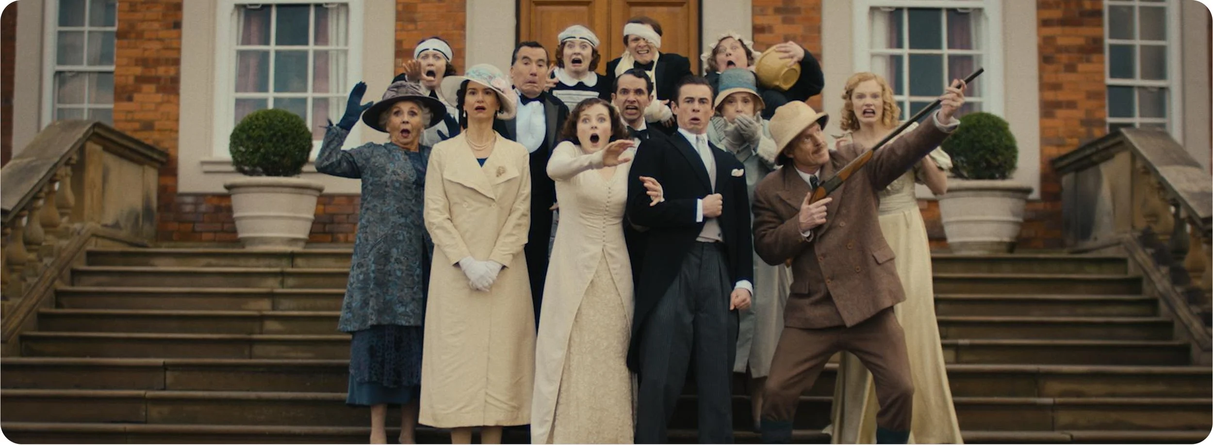 A group of people dressed in vintage clothing, standing on stairs in front of a brick building, reacting with surprise and shock. One man is holding a rifle, pointing it outward.