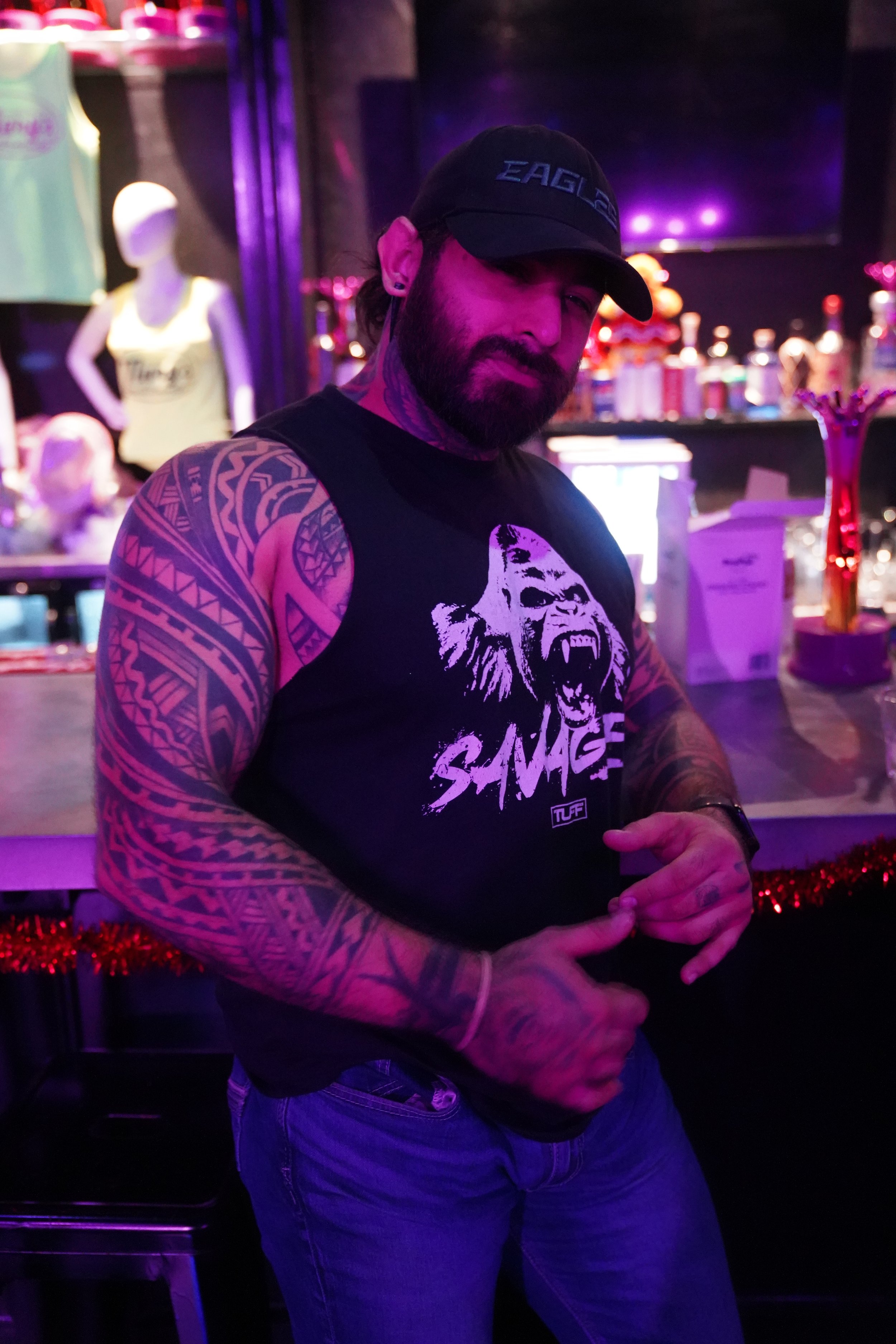 A man with tattoos on his arms, wearing a black sleeveless shirt with a graphic of a roaring lion and the word 'Savage,' a black cap, and blue jeans stands at a bar with purple and pink lighting.