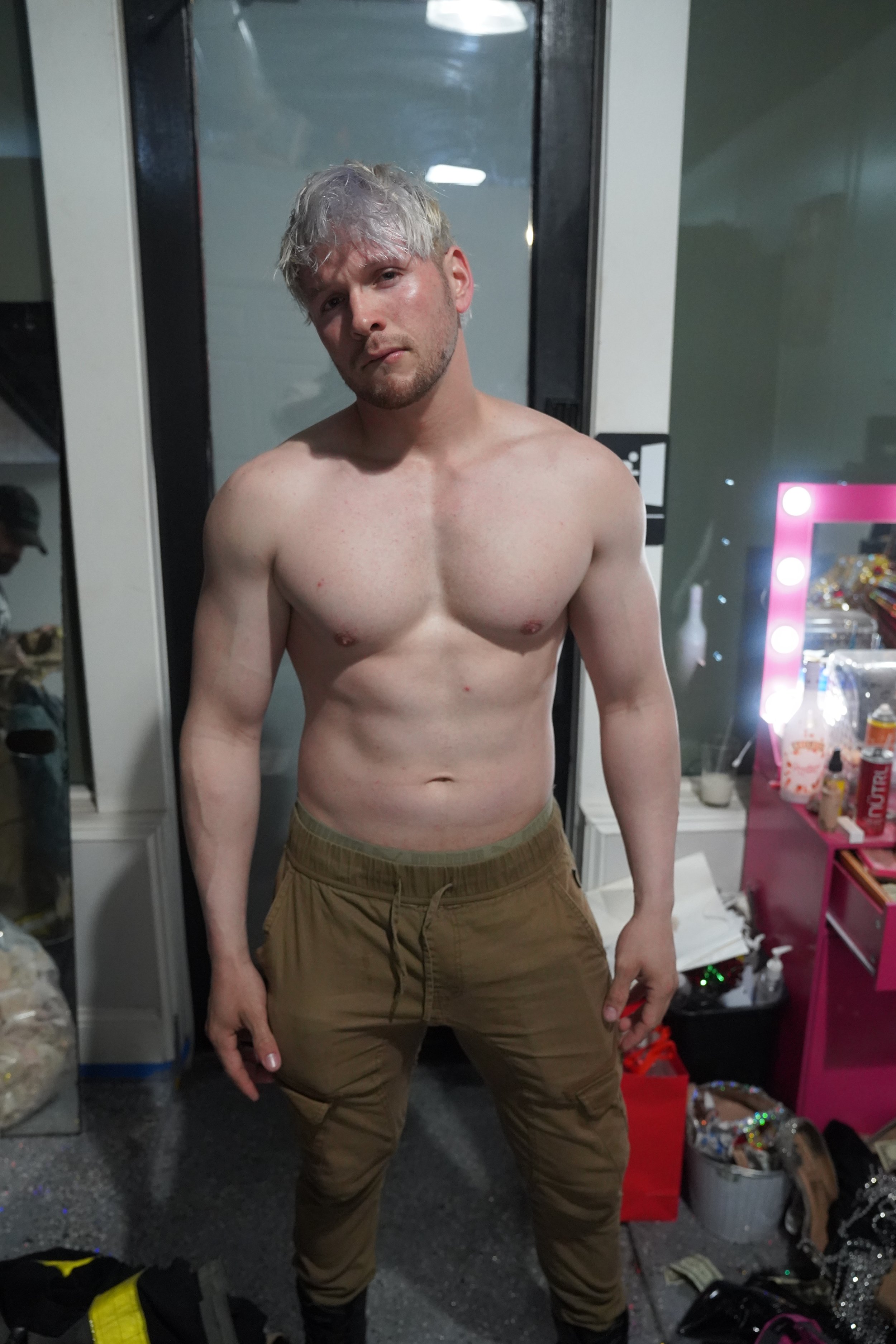 A shirtless young man with short, messy blond hair and a beard stands in front of a door, looking into the camera with a serious expression. He is wearing brown cargo pants and is positioned indoors near a dressing room or backstage area.