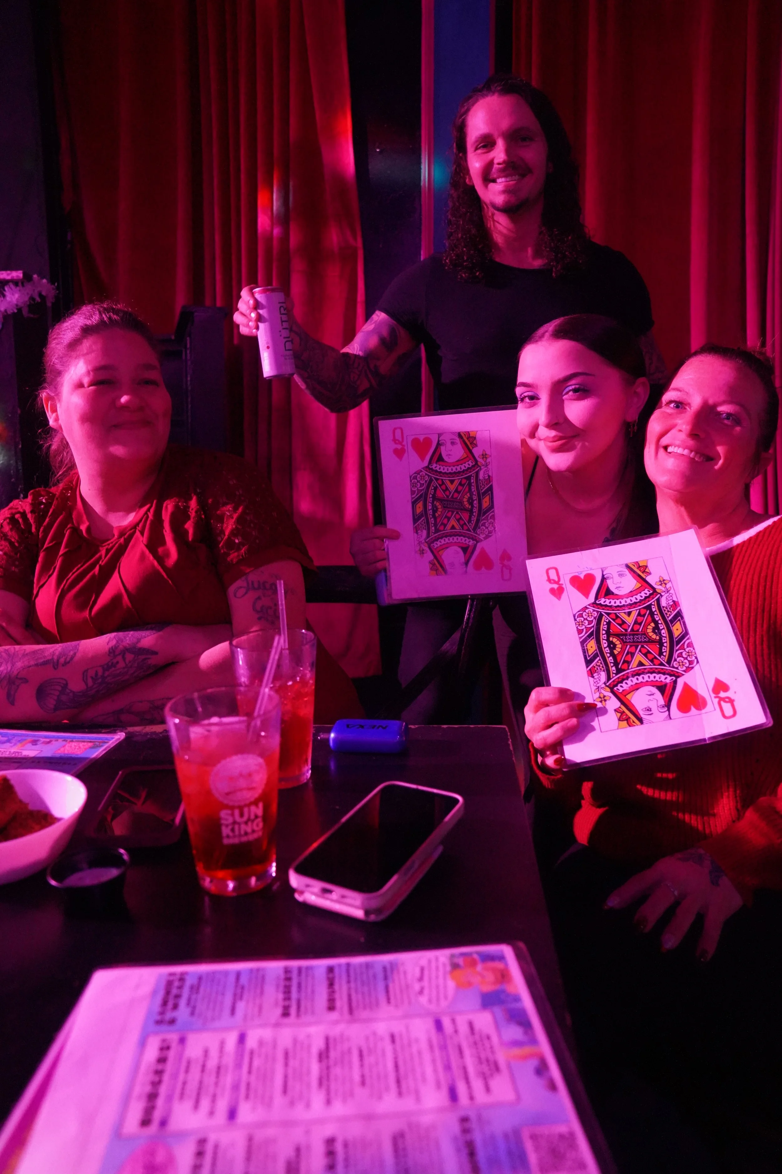 A group of four people at a party, smiling and holding Queens of hearts playing cards with a colorful queen playing card design. One person is standing behind the group, holding a drink and smiling. The table in front has drinks, a smartphone, a menu