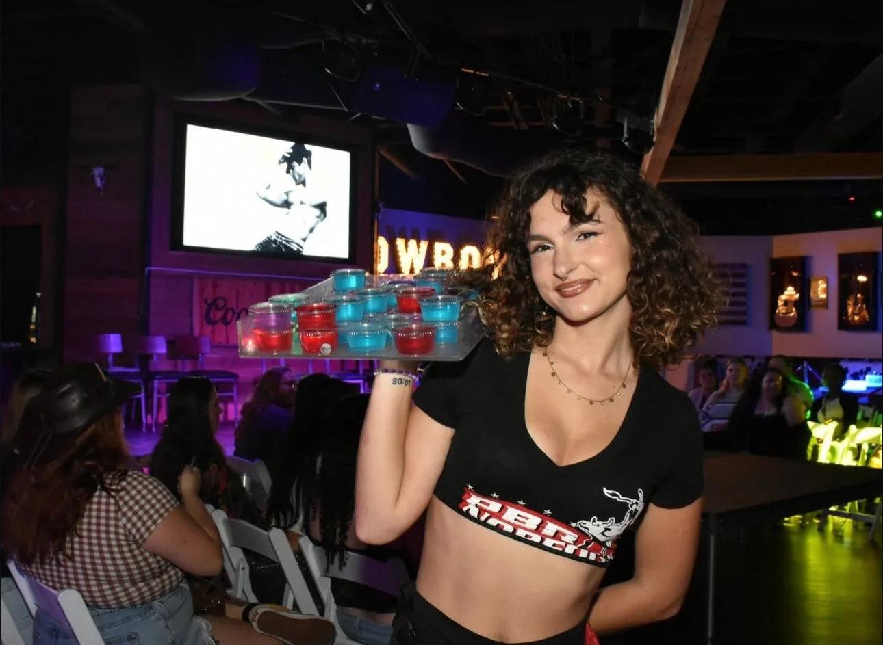 A woman with curly hair and a tie-dye crop top holds a tray of small, colorful candles at a dimly lit bar or club. She is smiling and looking at the camera, with a large screen and audience in the background.