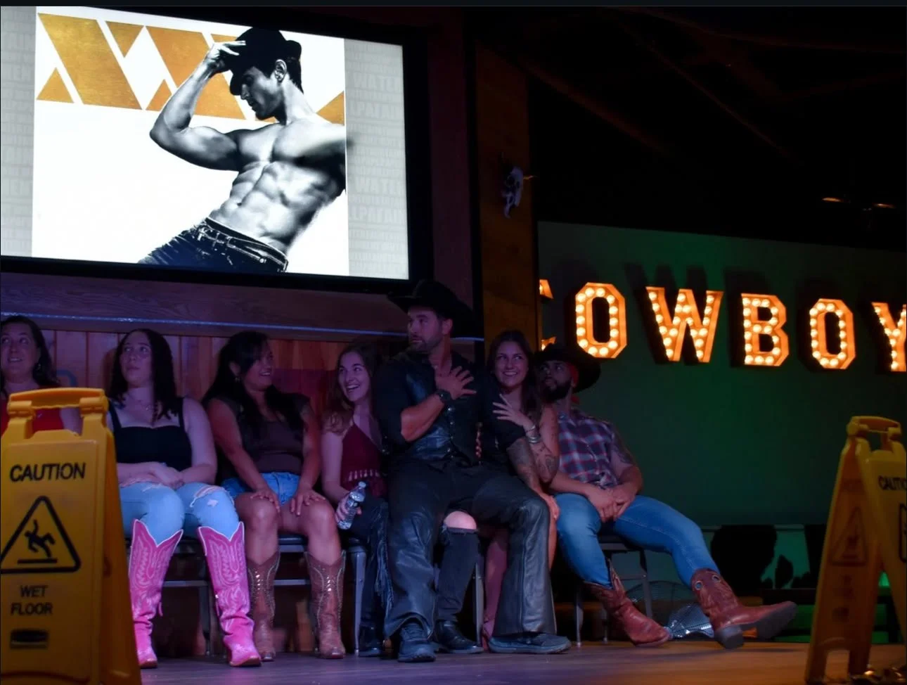 Group of six people sitting on a stage in front of a large screen displaying a black and white photo of a shirtless man with a muscular physique. Behind them, illuminated letters spell out "COWBOY." Two caution wet floor signs are at the bottom of th