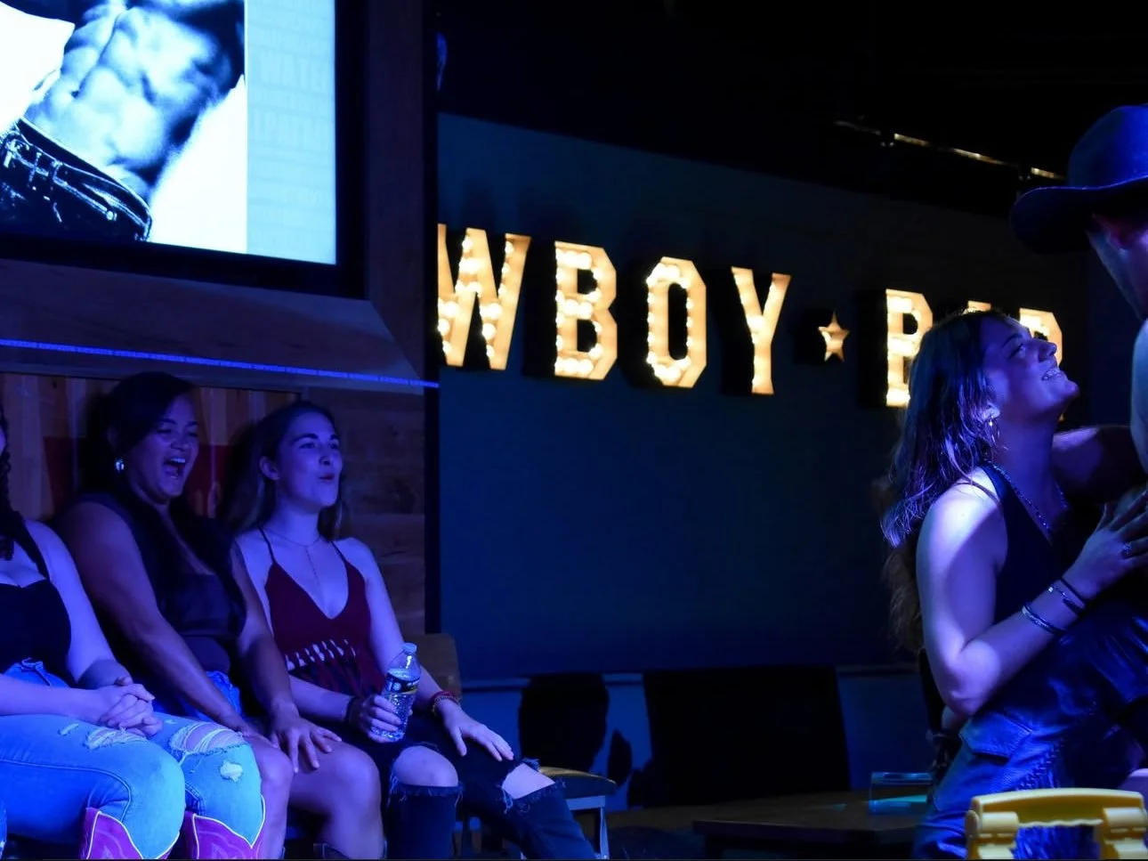 People sitting on a stage watching a performance, with illuminated letters behind them spelling 'WYBOY' in the background.
