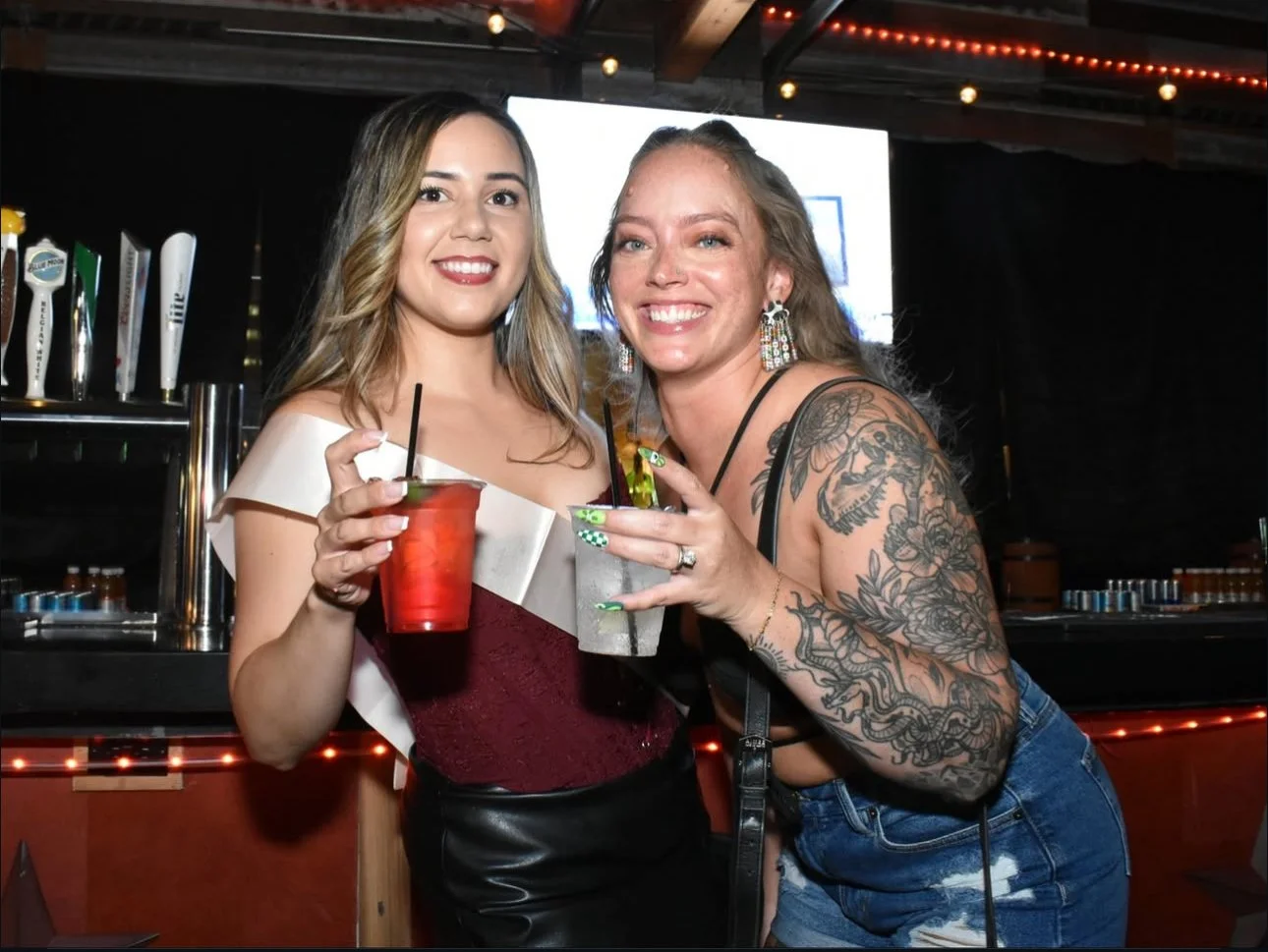Two women smiling and holding drinks at a bar, one with tattoos on her arm, night setting with bar equipment and string lights in the background.