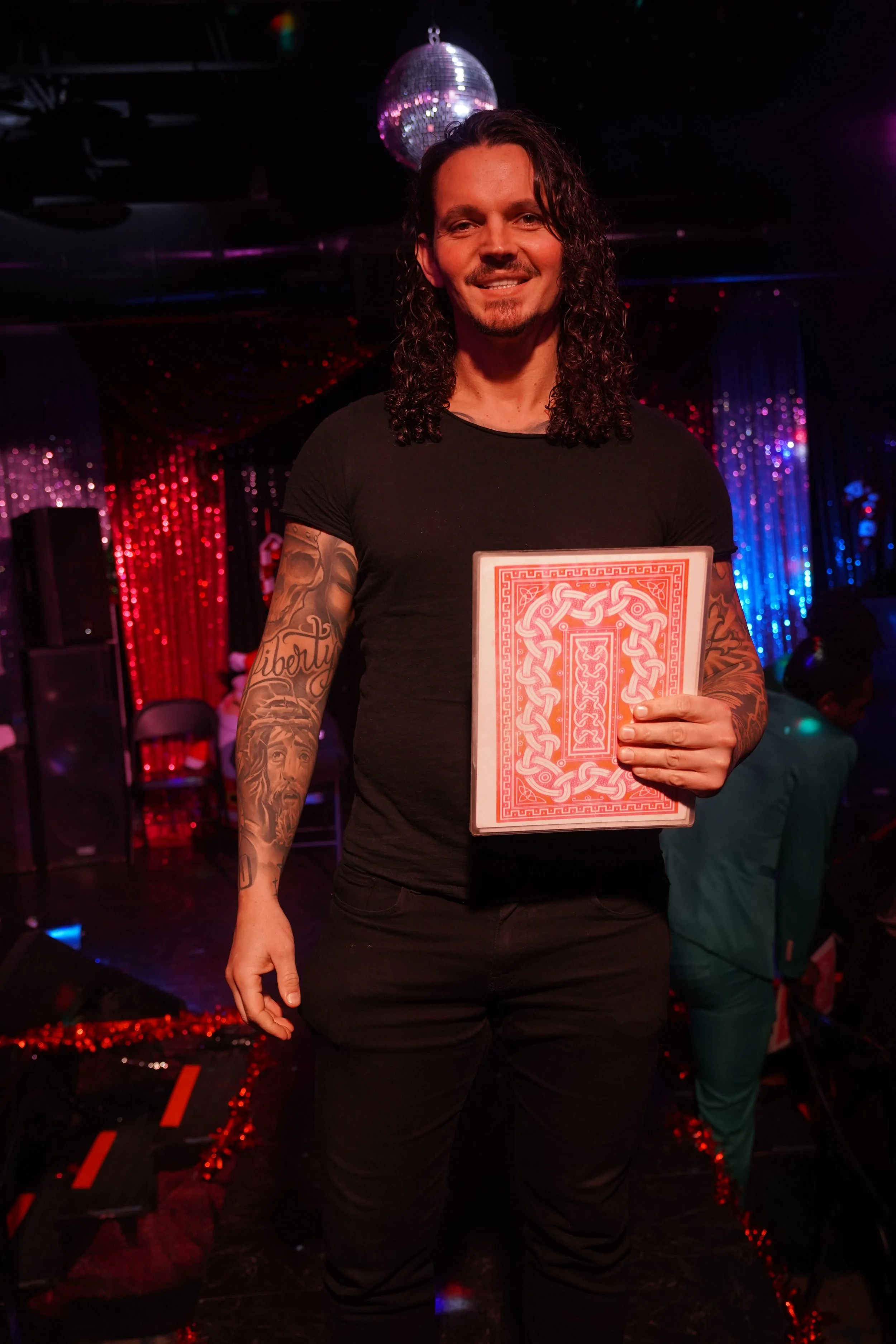 A man with long curly hair and tattoos on his arm, standing in a nightclub or bar, holding a red and white decorative object that resembles a large playing card or tarot card. The background features colorful shiny curtains and a disco ball.