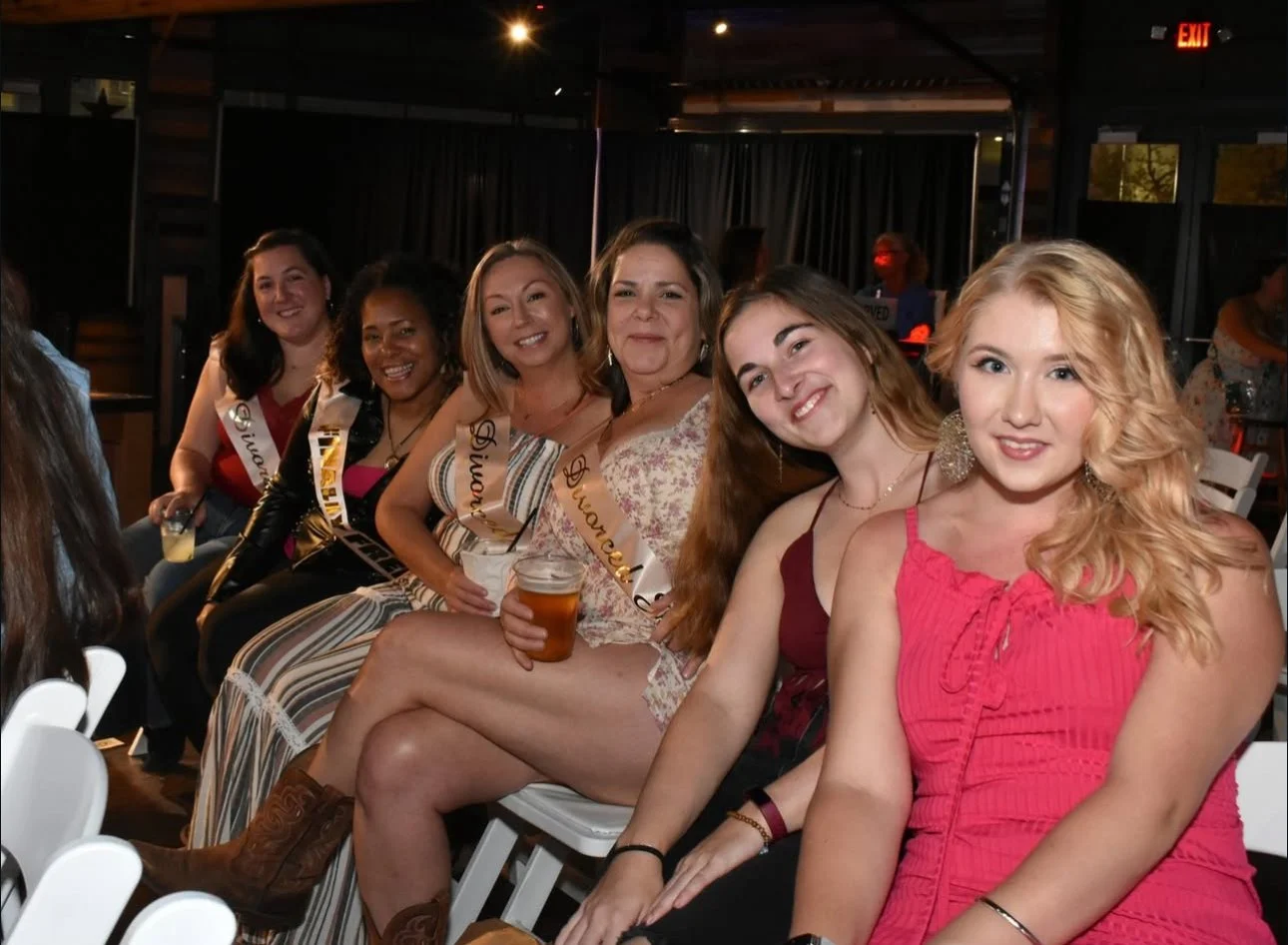 A group of six women sitting together at a social event, smiling for the camera. Five of them are wearing sashes indicating they might be pageant contestants or winners. They are holding drinks and dressed in casual to semi-formal attire, with some w