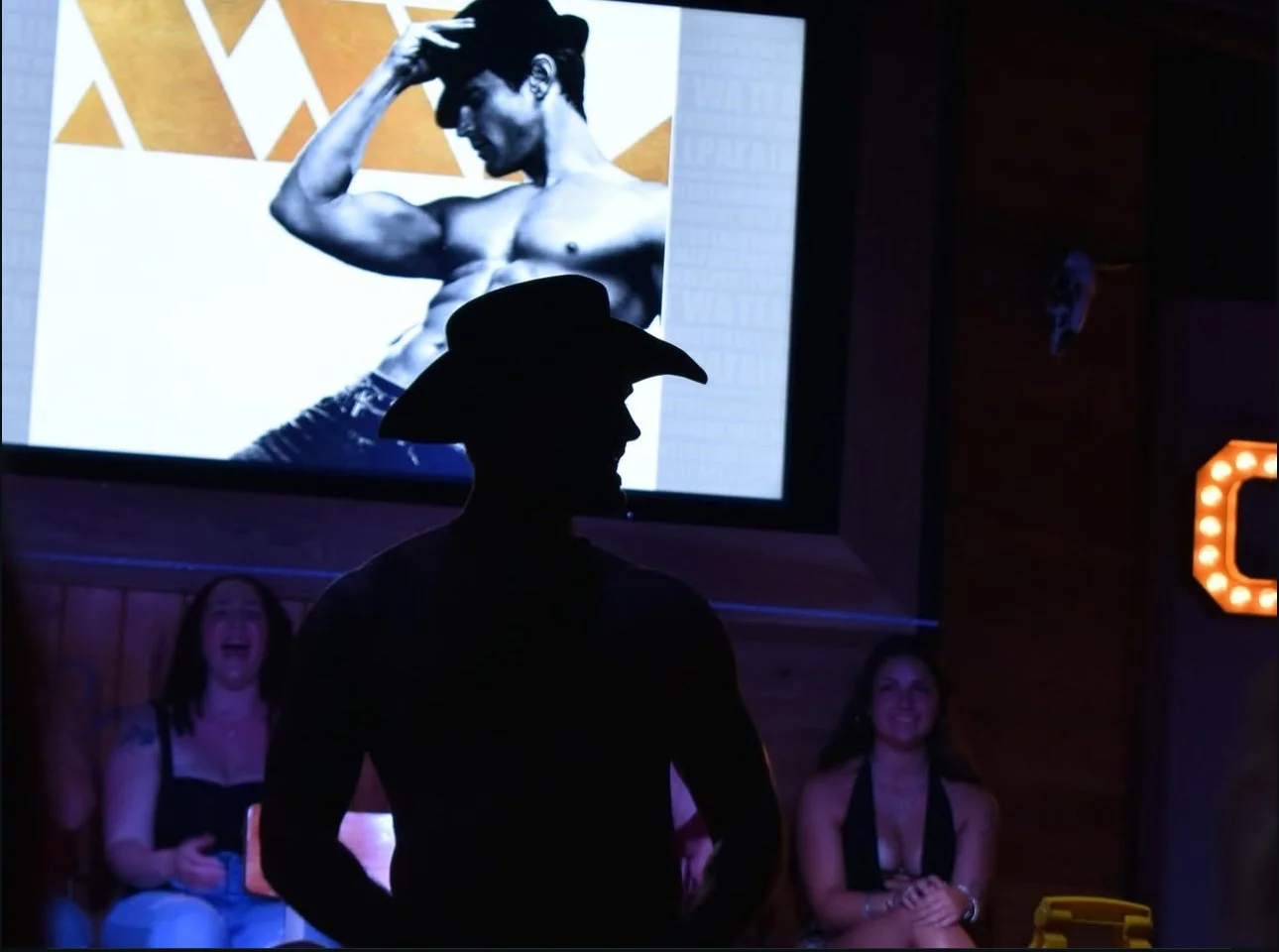A man wearing a cowboy hat in silhouette, standing in front of a large screen with a black-and-white photo of a shirtless man flexing his arm. Two women in the background are sitting and smiling.