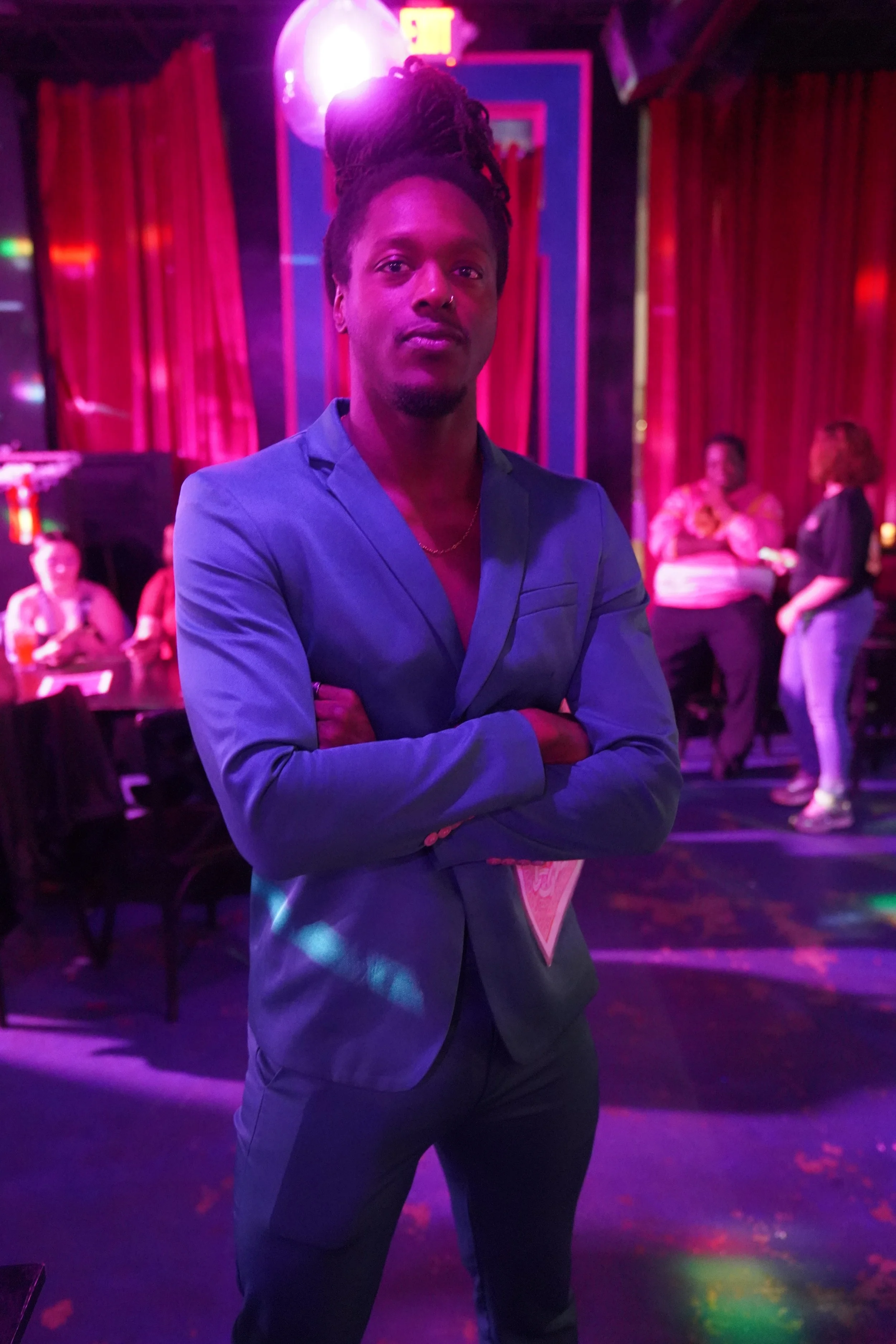 A man in a blue suit with dreadlocks standing with arms crossed in a dimly lit, colorful nightlife venue, with a bar and other guests in the background.