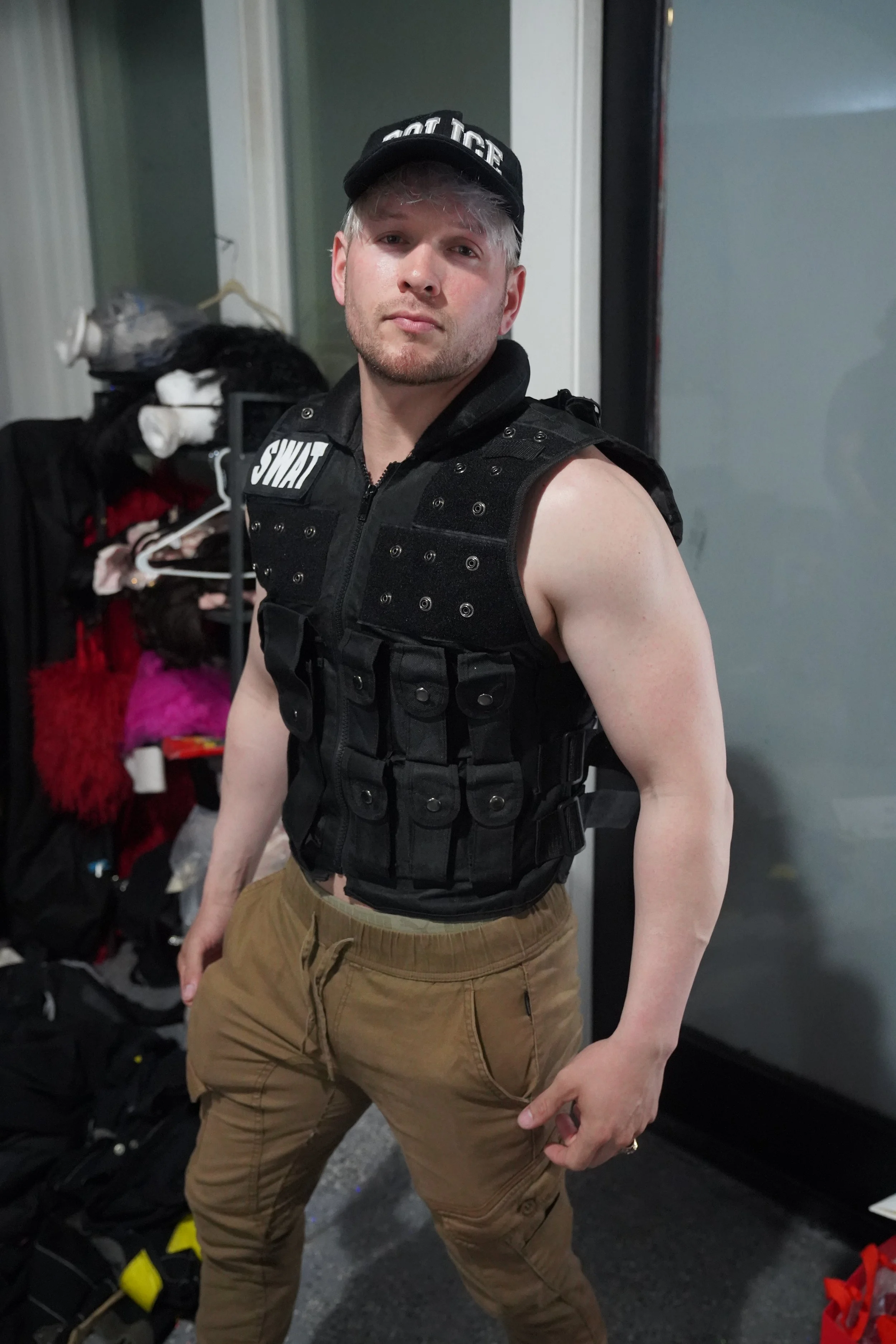 A young man wearing a black SWAT tactical vest, a black cap, and tan pants, standing indoors with a confident expression, near a closet or storage area with clothes and accessories.