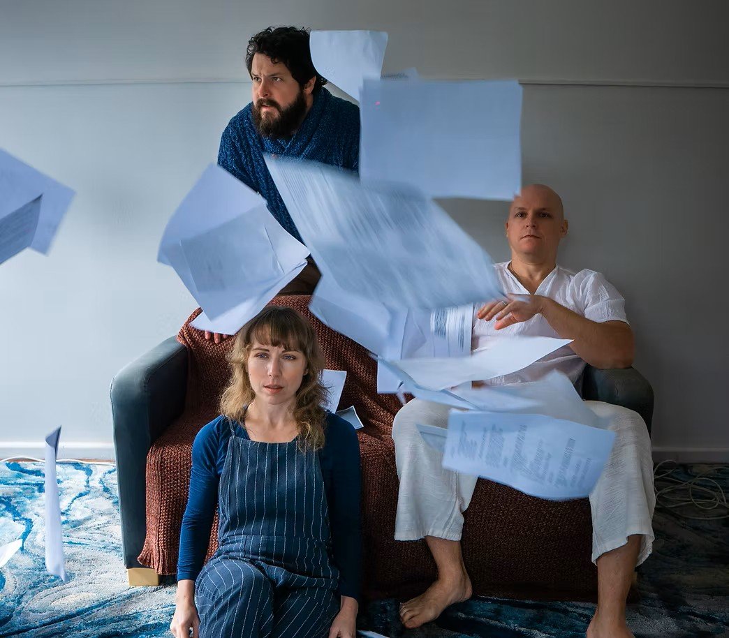 Three people sitting on a couch with papers flying around them. A woman with brown hair wearing a blue dress with a striped apron is sitting on the floor in front of the couch. A man with a bald head wearing white pajamas is sitting on the right side of the couch. Another man with dark curly hair and beard wearing a blue robe is peeking over the top of the couch.
