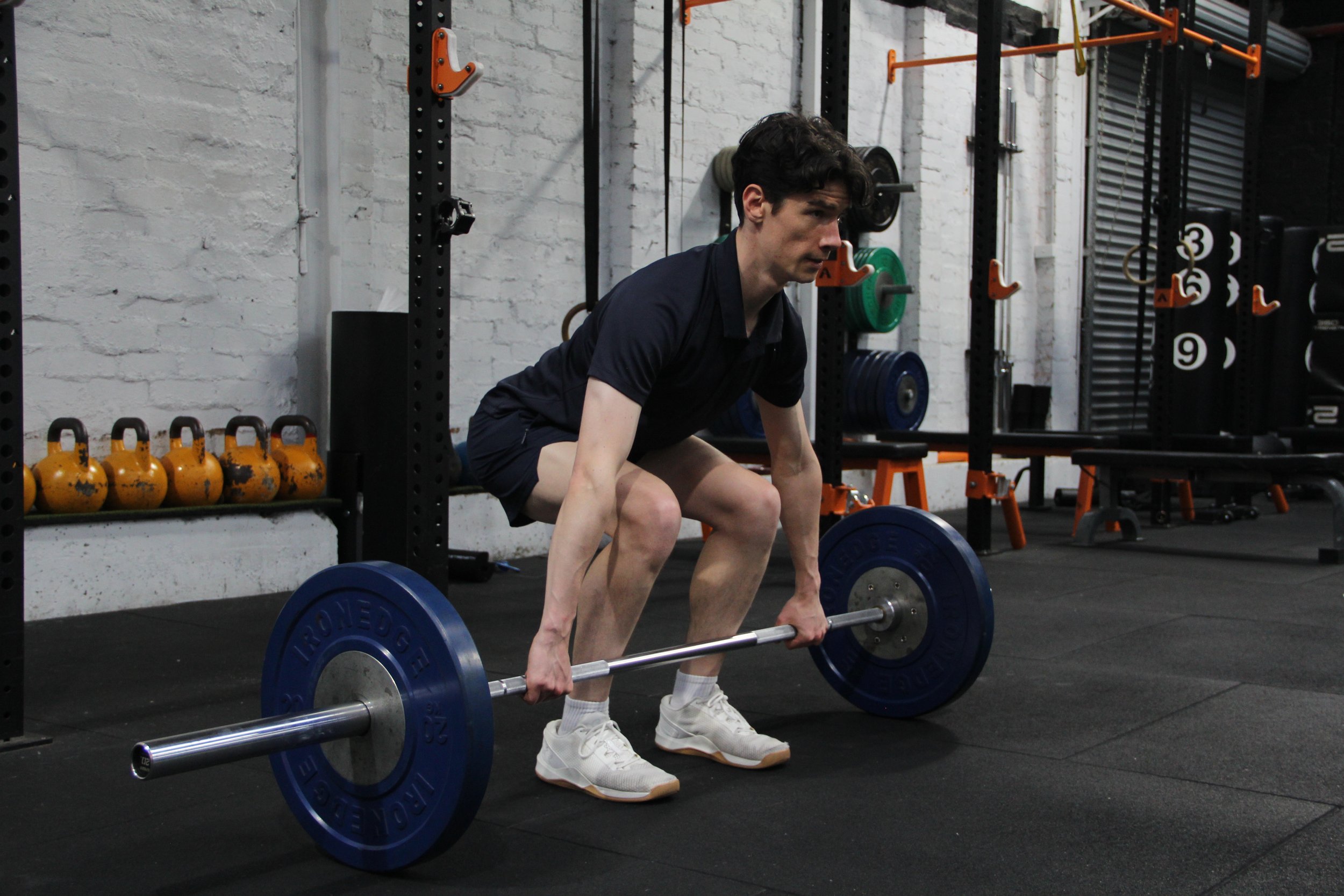 A man with dark hair wearing a dark t-shirt, shorts, white socks, and sneakers is preparing to lift a barbell with blue weight plates at a gym. The gym has a white brick wall, various weight plates, kettlebells, and exercise equipment in the background.