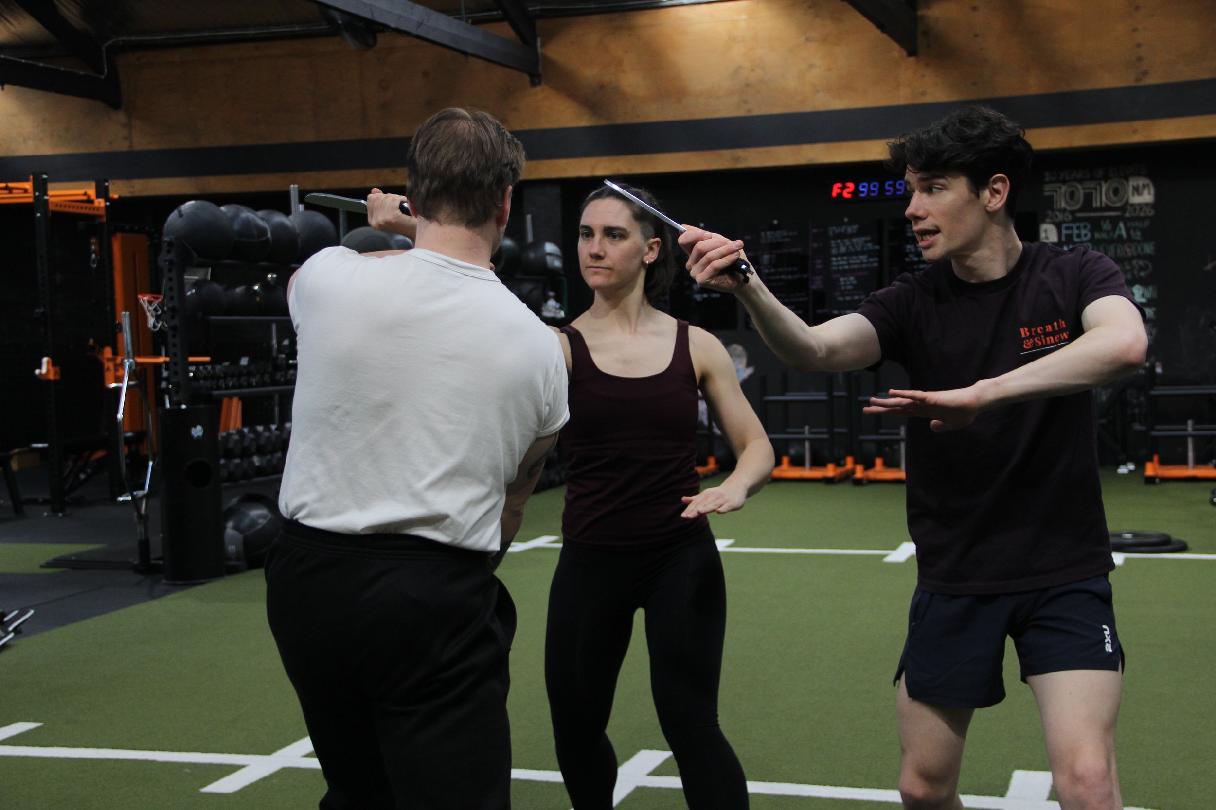 Three people training in a gym. A man with short hair in a white shirt, a woman in a maroon tank top, and a young man with dark curly hair in a black shirt. The young man is holding a stick, and the man with short hair is holding a kettlebell, possibly for self-defense training.