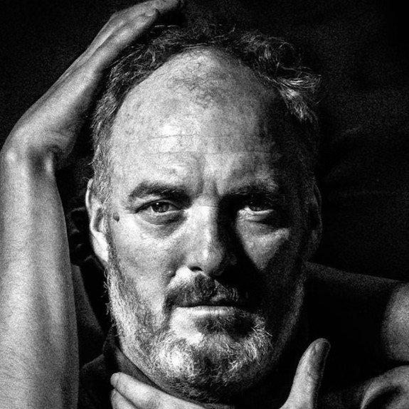 Close-up black and white photo of a man with a beard and mustache, looking directly at the camera, with one hand resting on his head and the other near his chin.