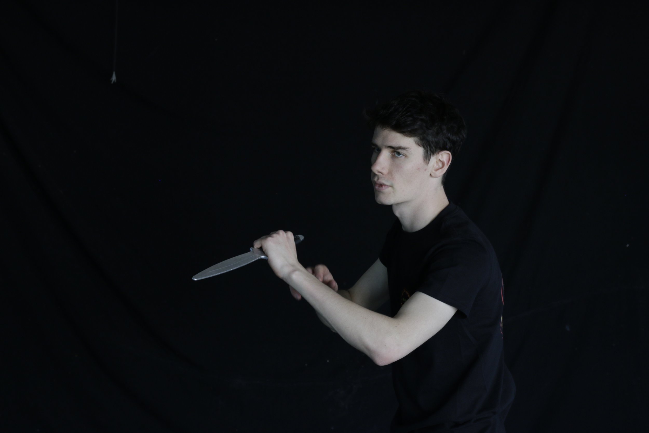 A young man with dark hair and fair skin, wearing a black t-shirt, holding a large kitchen knife with his right hand, standing against a black background.