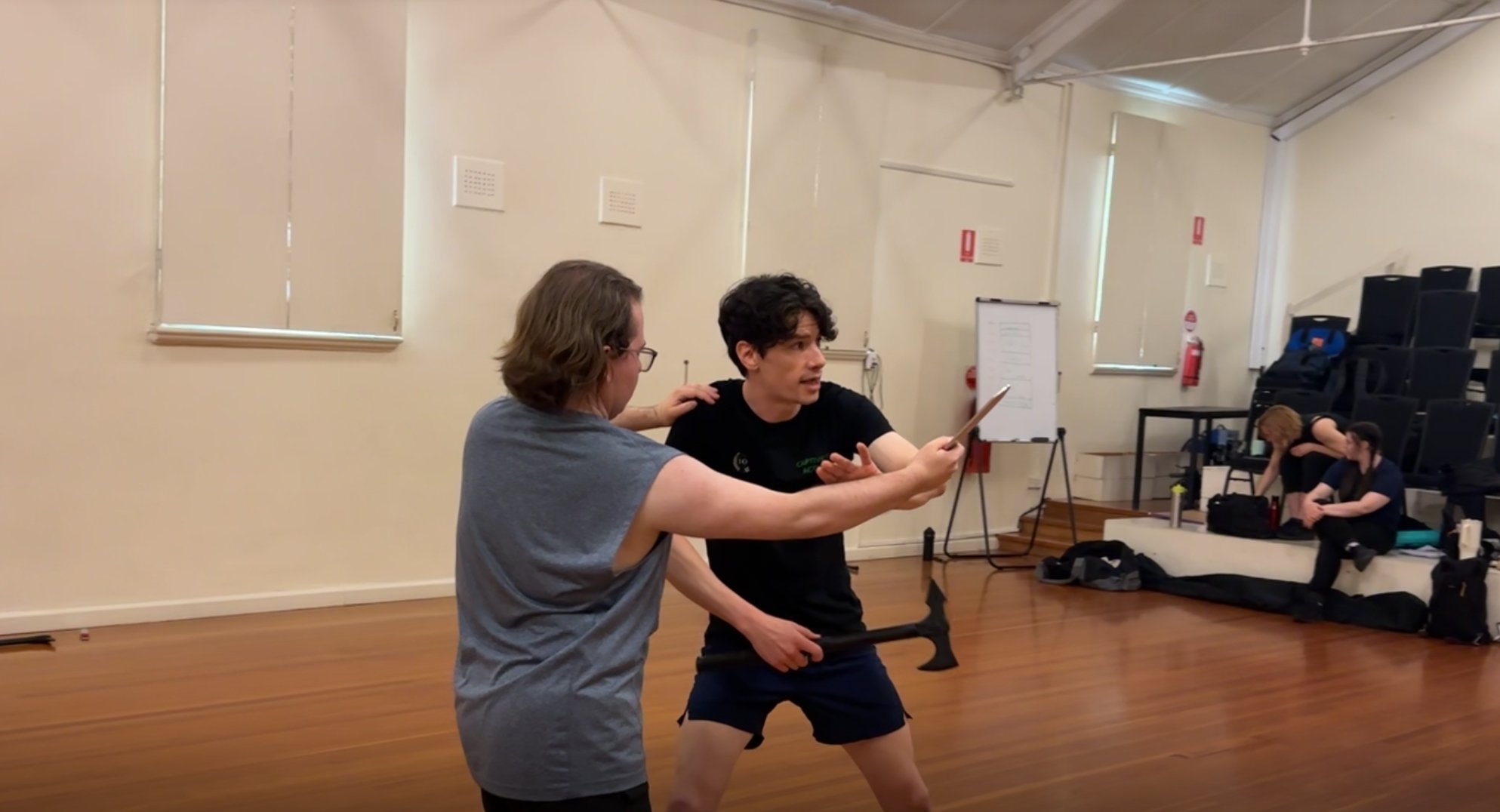 Two men practicing martial arts techniques in a studio, with three women sitting on mats in the background, attending a class or training session.