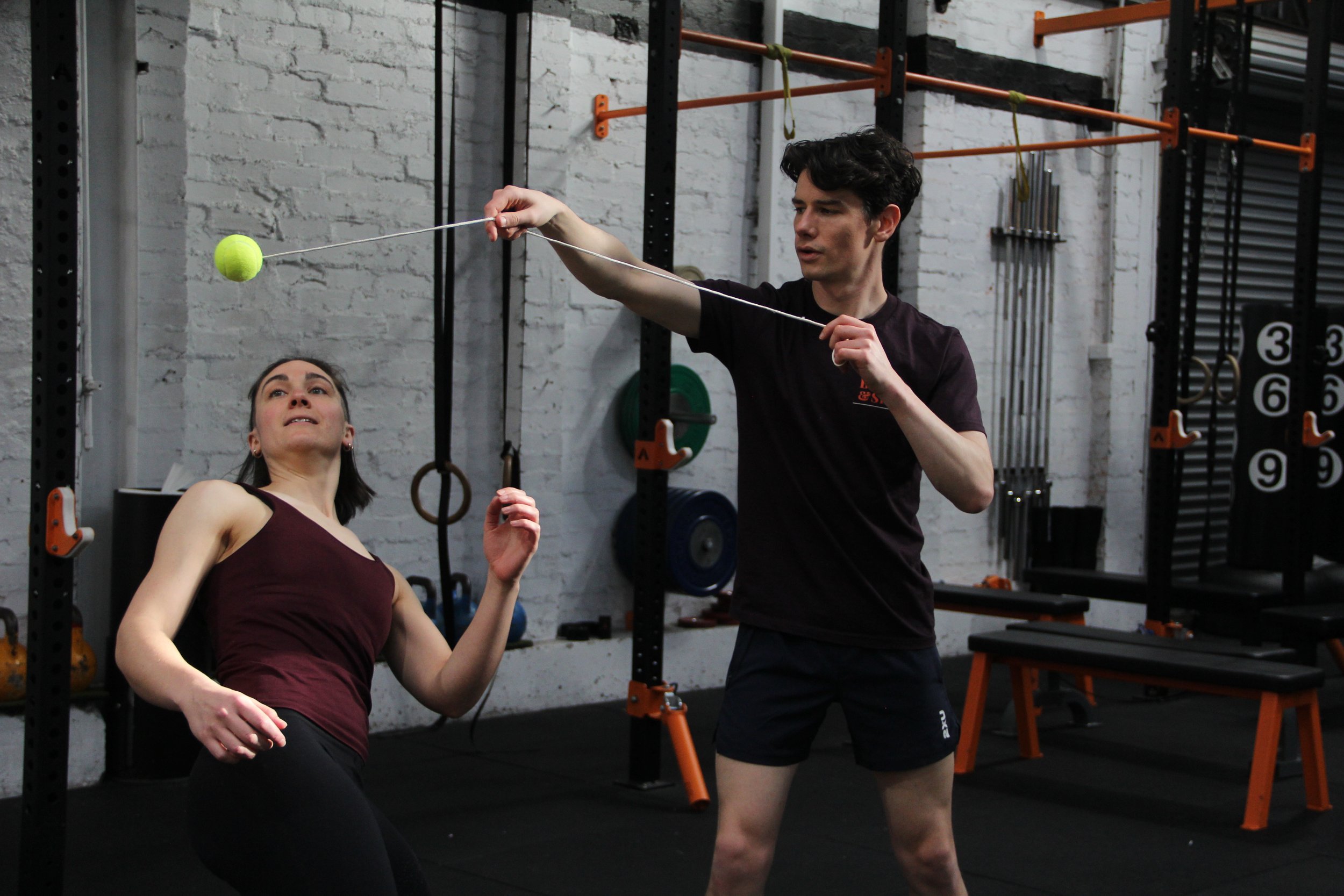 A man and a woman training with a tennis ball in a gym. The woman is sitting and looking up at the tennis ball; the man is standing and holding a string connected to the ball.