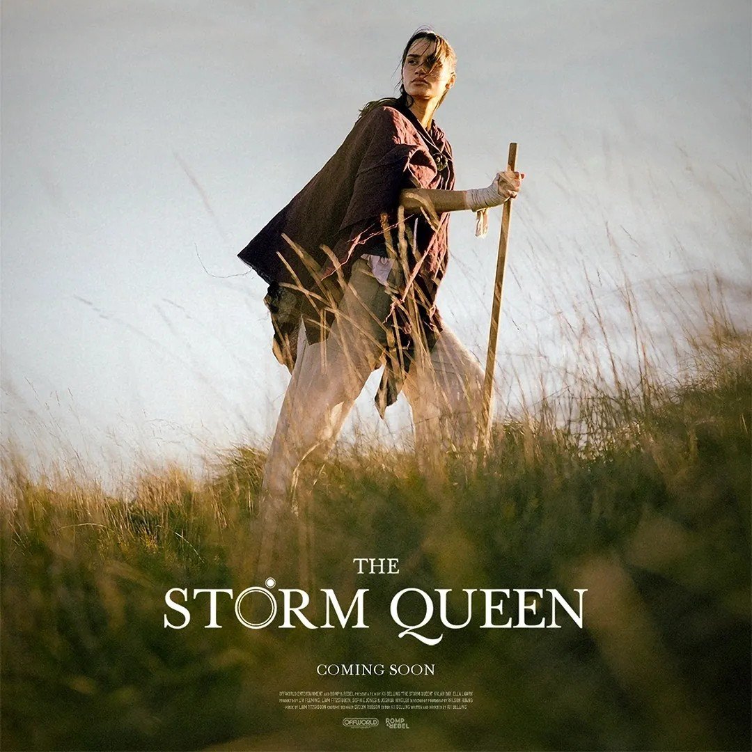 A woman holding a stick standing in a grassy field, with a dramatic sky in the background, promotional poster for 'The Storm Queen'.