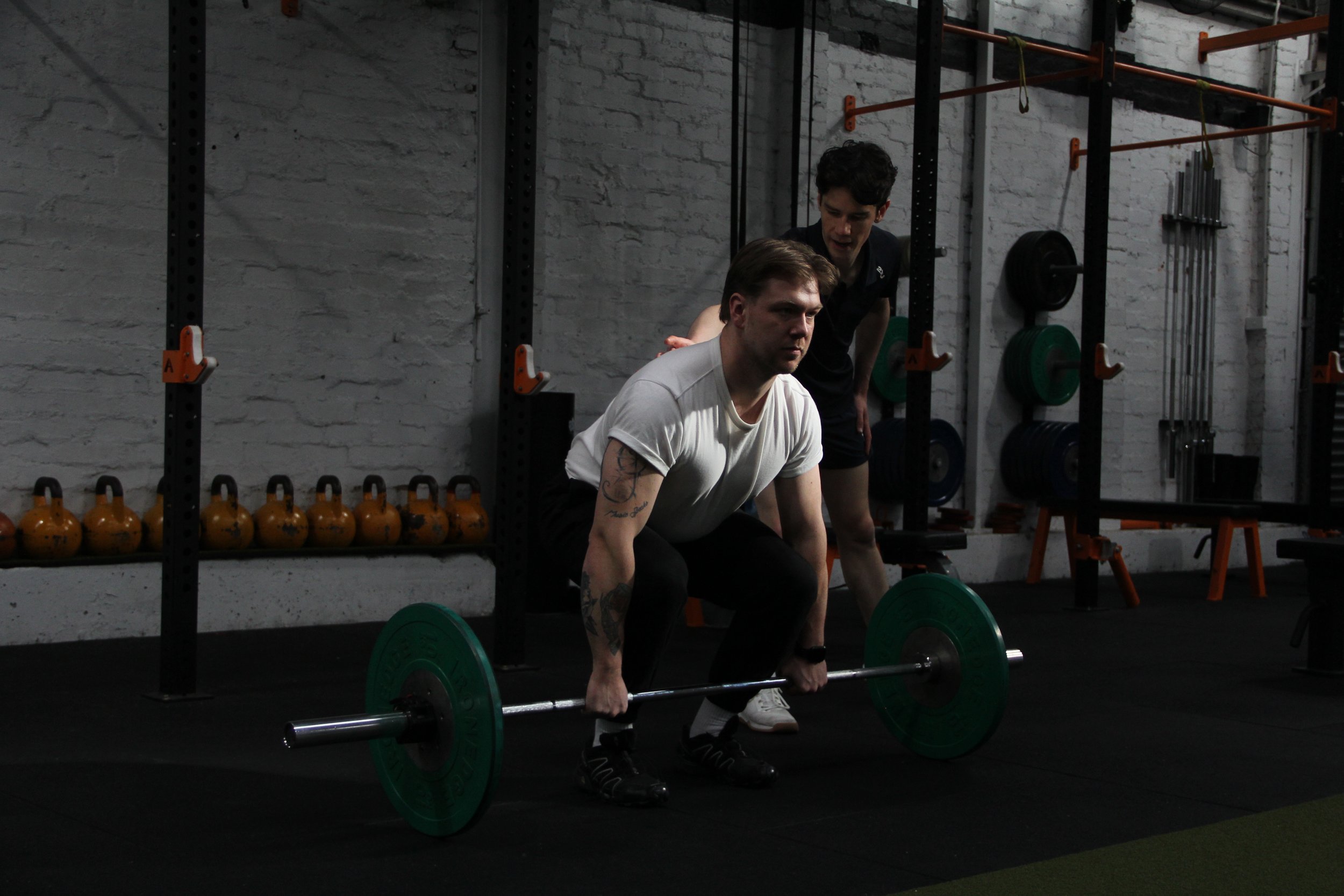 Two men are in a gym, one is lifting a barbell with green weights while the other is standing beside him, coaching. The gym has a brick wall with weightlifting equipment and kettlebells.