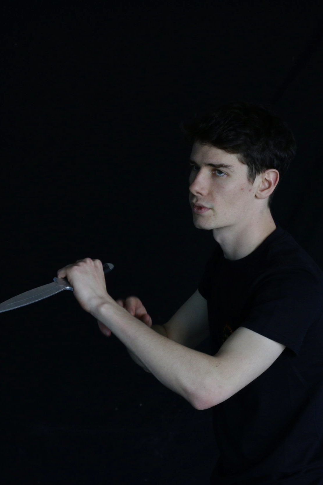 A young man with dark hair wearing a black t-shirt holding a sword in a dark room.