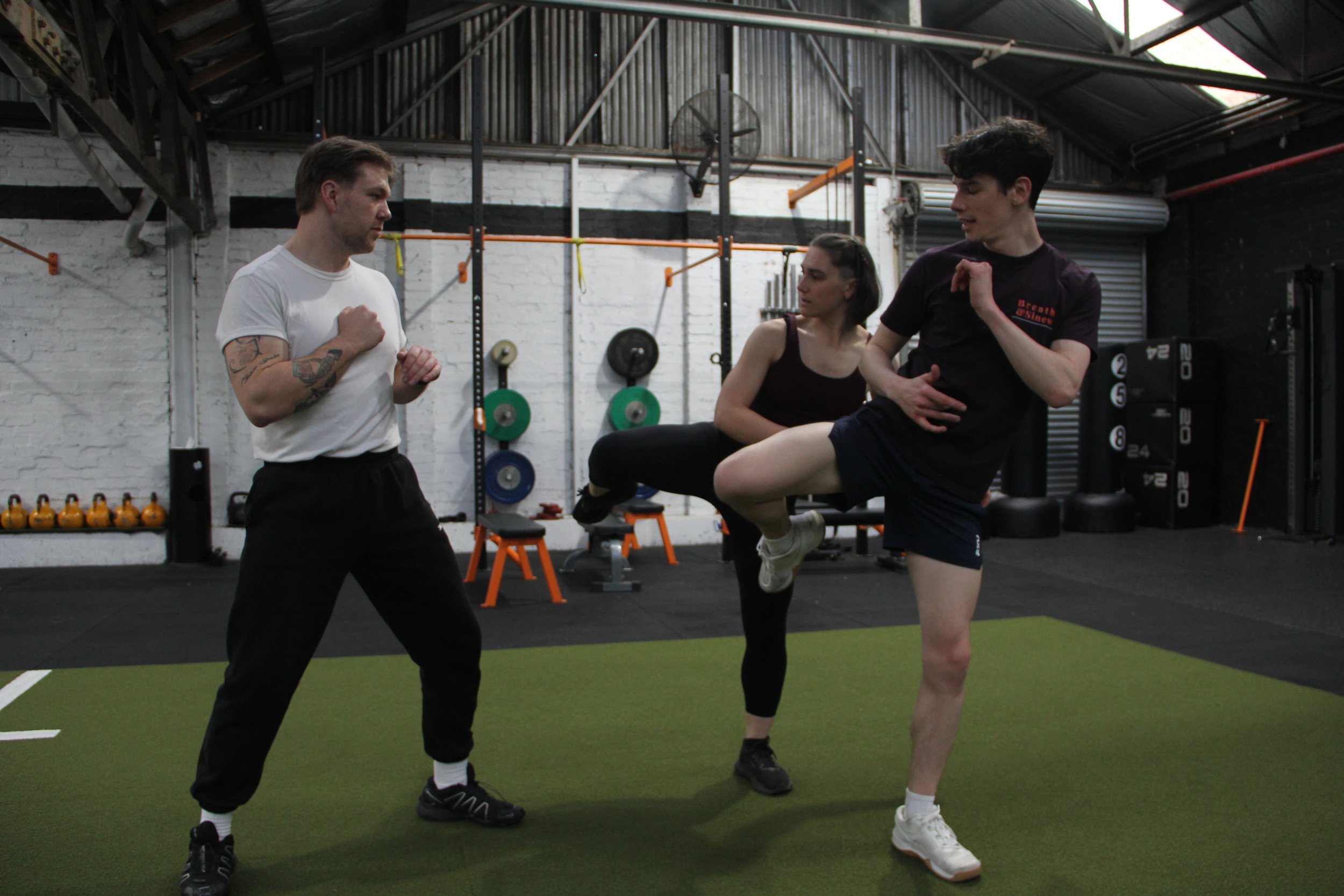Three people exercising in a gym, two men and one woman, with gym equipment visible in the background, including weights, barbells, and medicine balls.