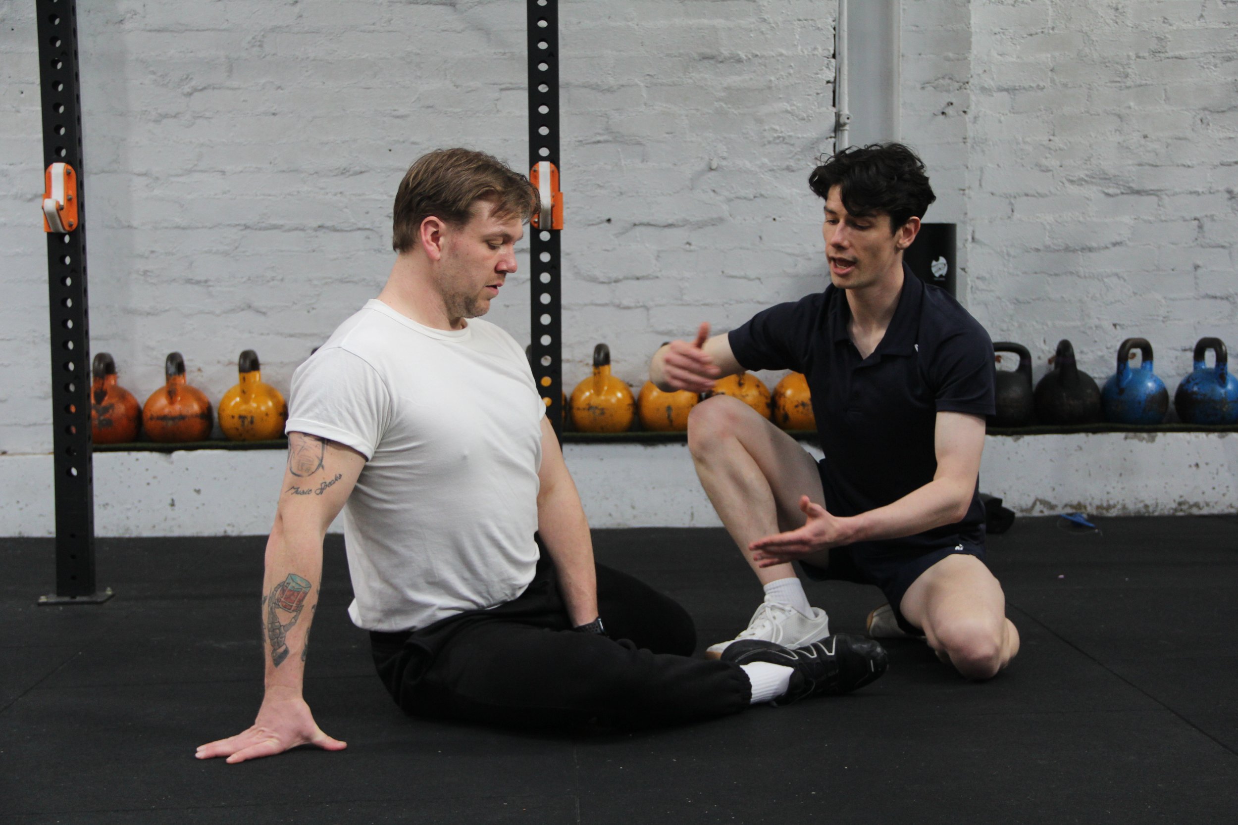 Two men training with one sitting on the floor and the other coaching in a gym with kettlebells in the background.