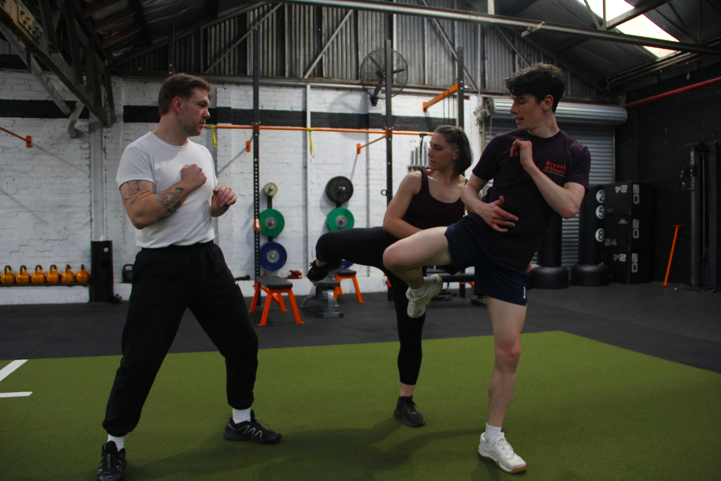 Three people in a gym practicing martial arts or self-defense techniques. One person is standing with fists up, while two others are engaged in a training exercise involving a leg sweep. The gym has black and white walls with fitness equipment visible in the background.