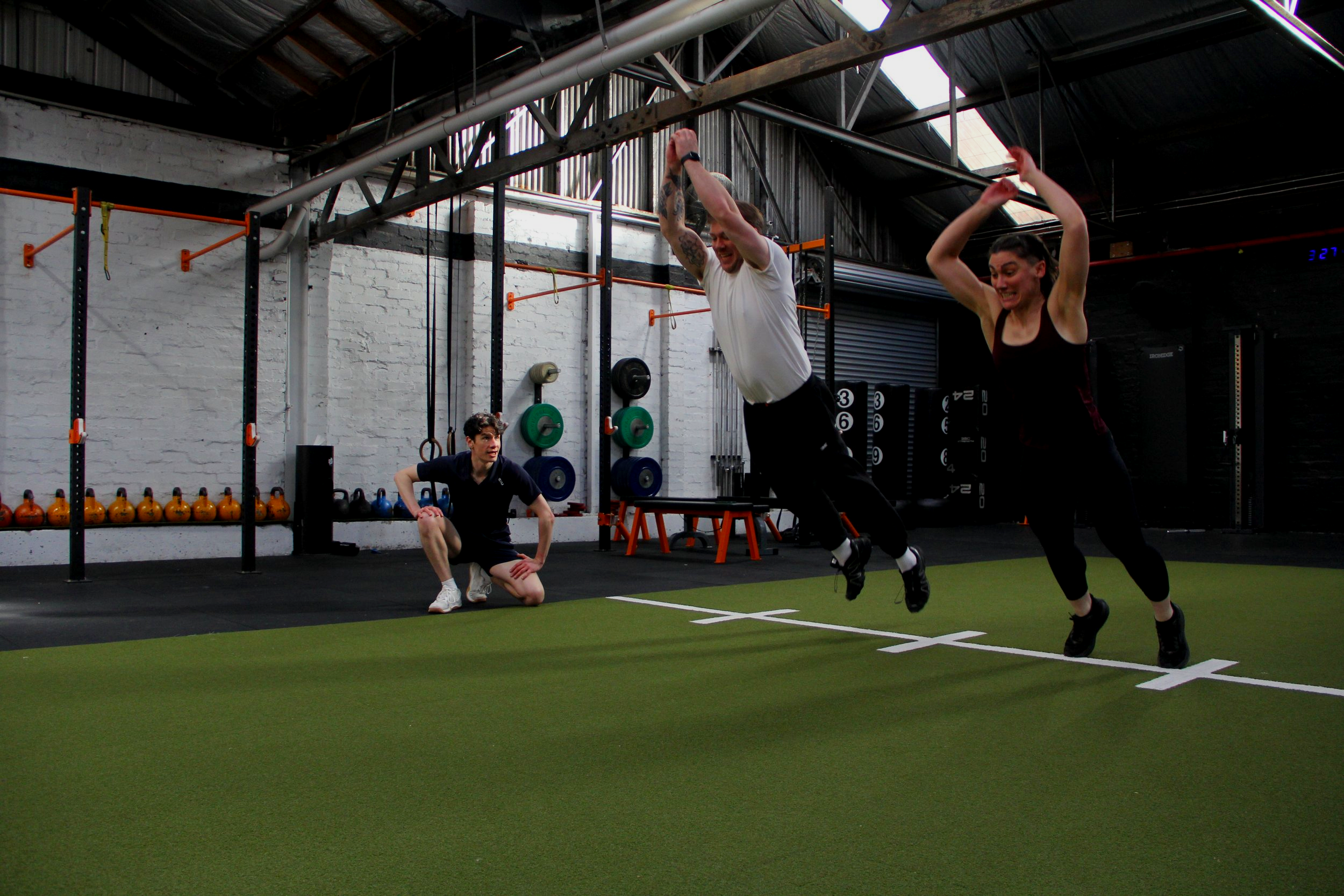 Two women and a man exercising in a gym with artificial turf, weightlifting equipment, and exposed pipes in the background.