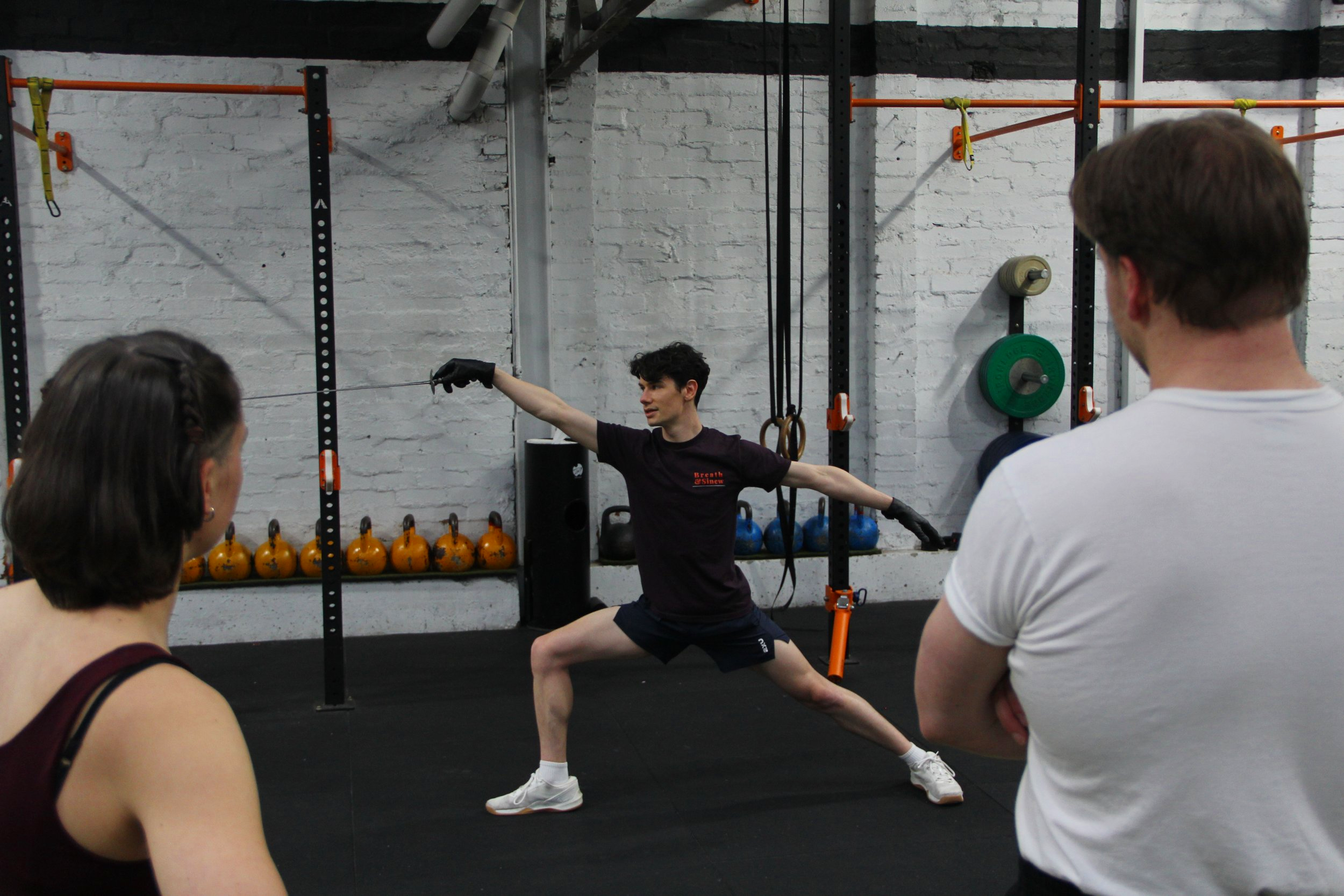 A group fitness class with four participants and an instructor demonstrating a fencing posture in a gym with black flooring, white brick walls, and workout equipment in the background.