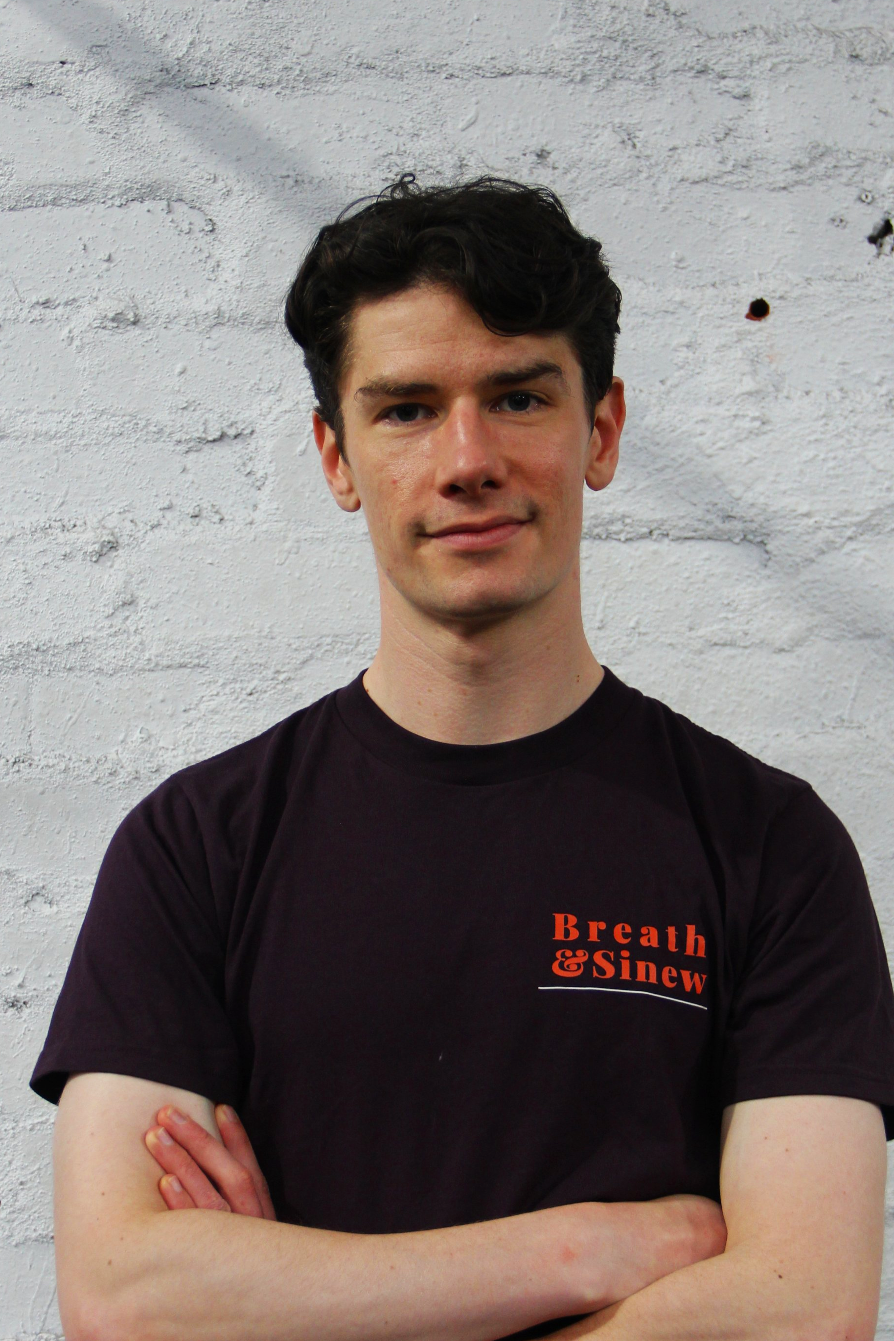A young man with curly dark hair and light skin, standing against a white brick wall, wearing a black t-shirt with orange text that reads 'Breath & Sinew'. He has a slight smile, arms crossed, and is looking directly at the camera.