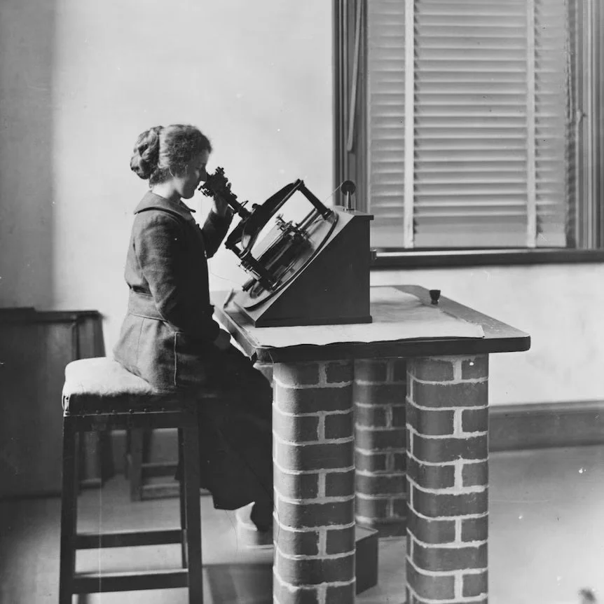 A young girl in vintage clothing sits on a stool, looking into a scientific microscope positioned on a desk with a brick base, in a room with a window and blinds.