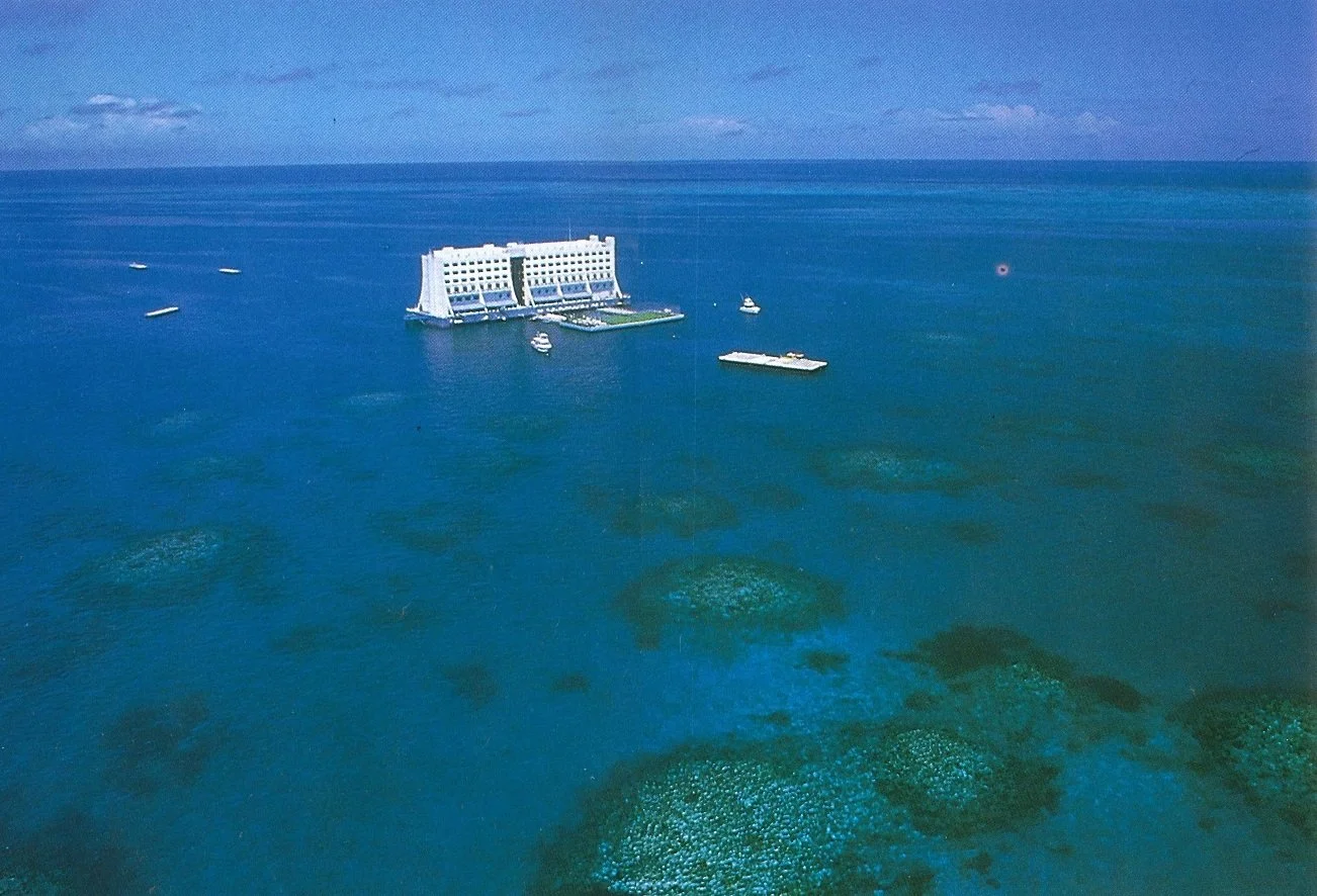 Aerial view of a large hotel building surrounded by boats in the ocean with coral reefs visible beneath the water.