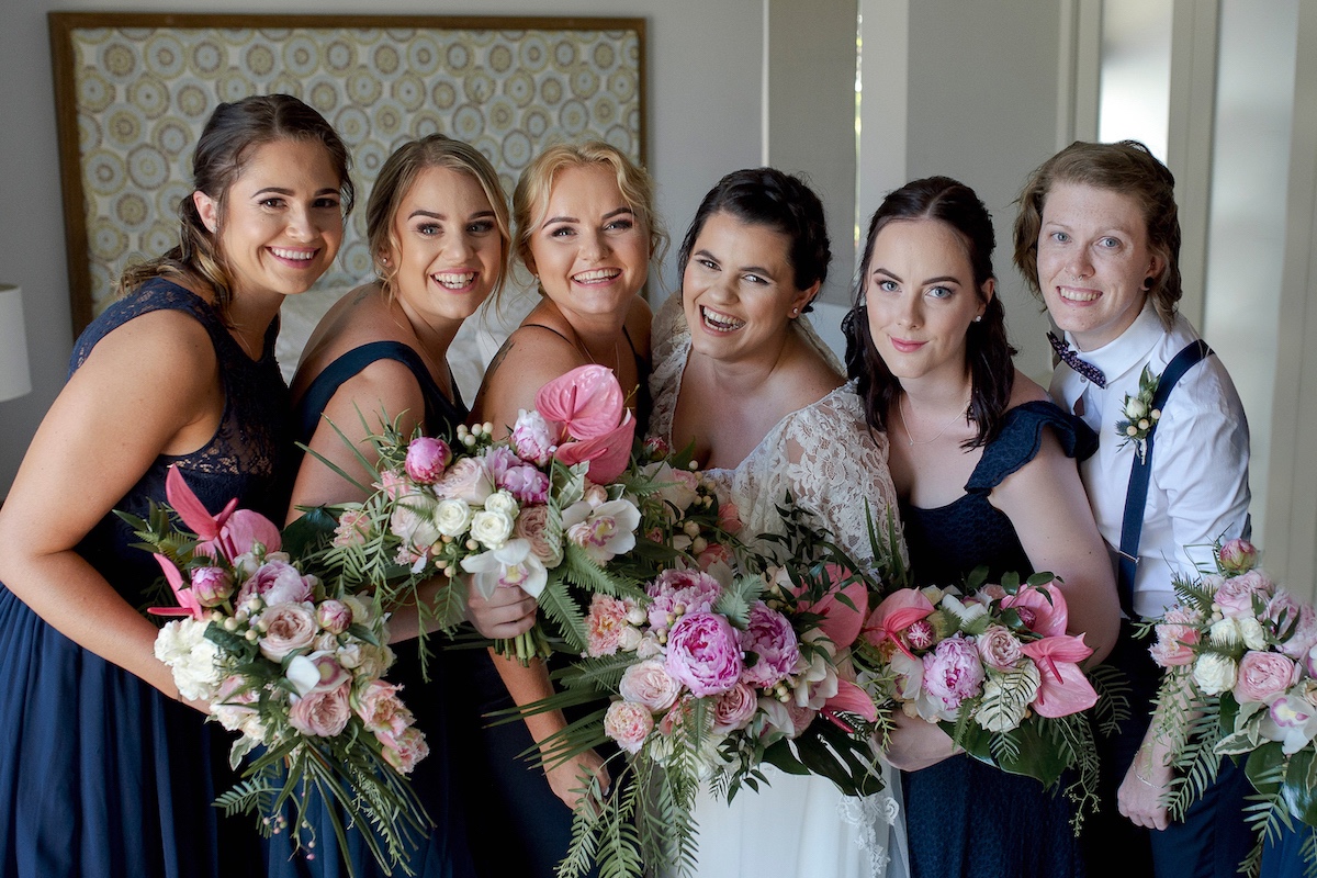 Bride and bridesmaids in navy dresses with pink peony and tropical bouquets.