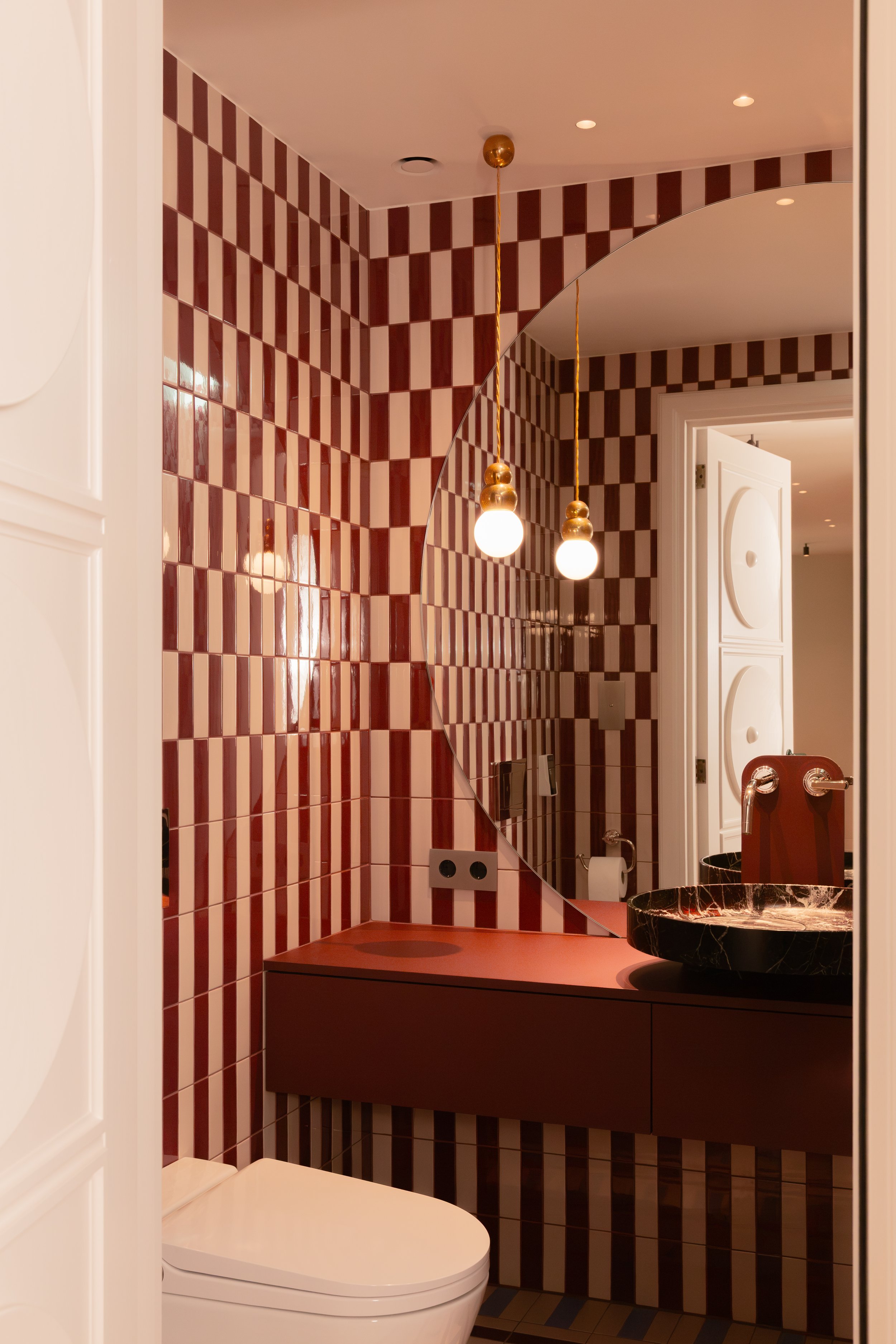 10. Burgundy powder room: Private restroom featuring contrasting bordeaux and white Quintessenza Ceramiche Argblock patterned tiles, a semicircle mirror, a hanging gold lamp, a marble basin, and Watermark collection accessories.