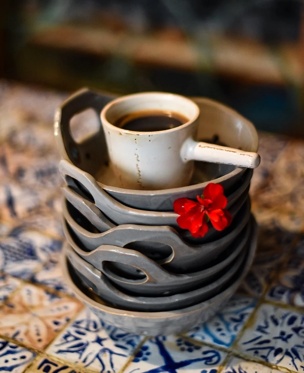 Stack of gray and white coffee cups on a patterned tile surface, with one cup filled with coffee and a red flower placed on the cups.
